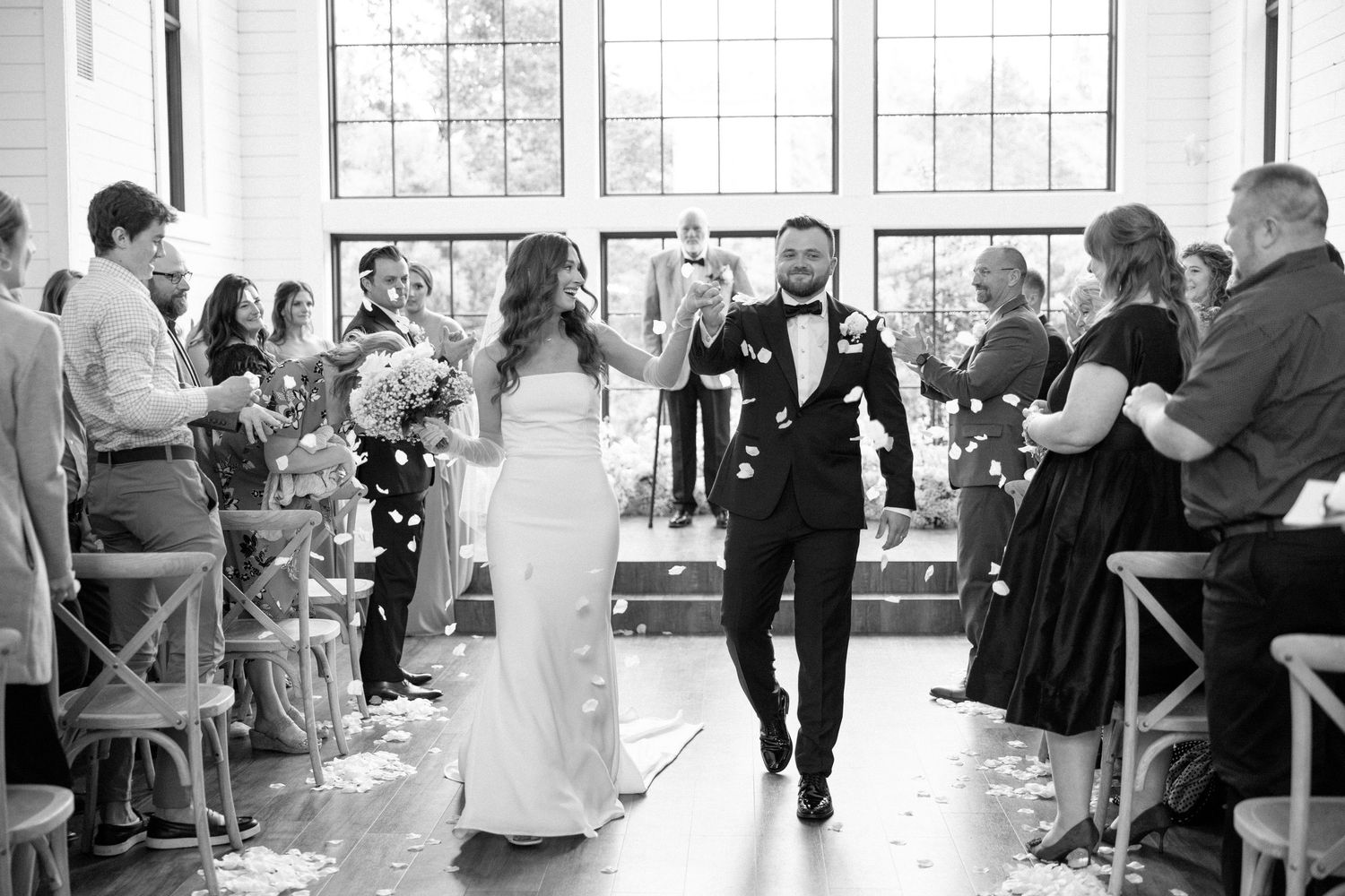 A joyful couple walks down the aisle while guests throw confetti during an indoor wedding ceremony.