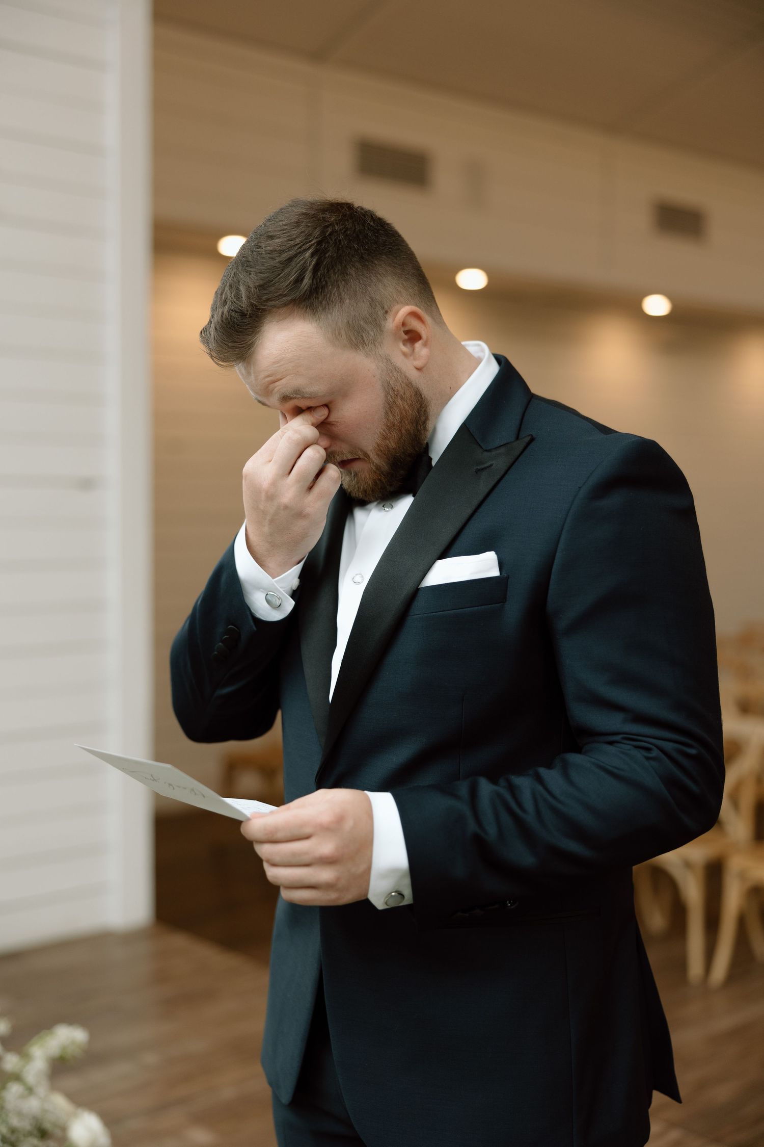 Man in black suit reads emotional letter while wiping tears during wedding preparations.