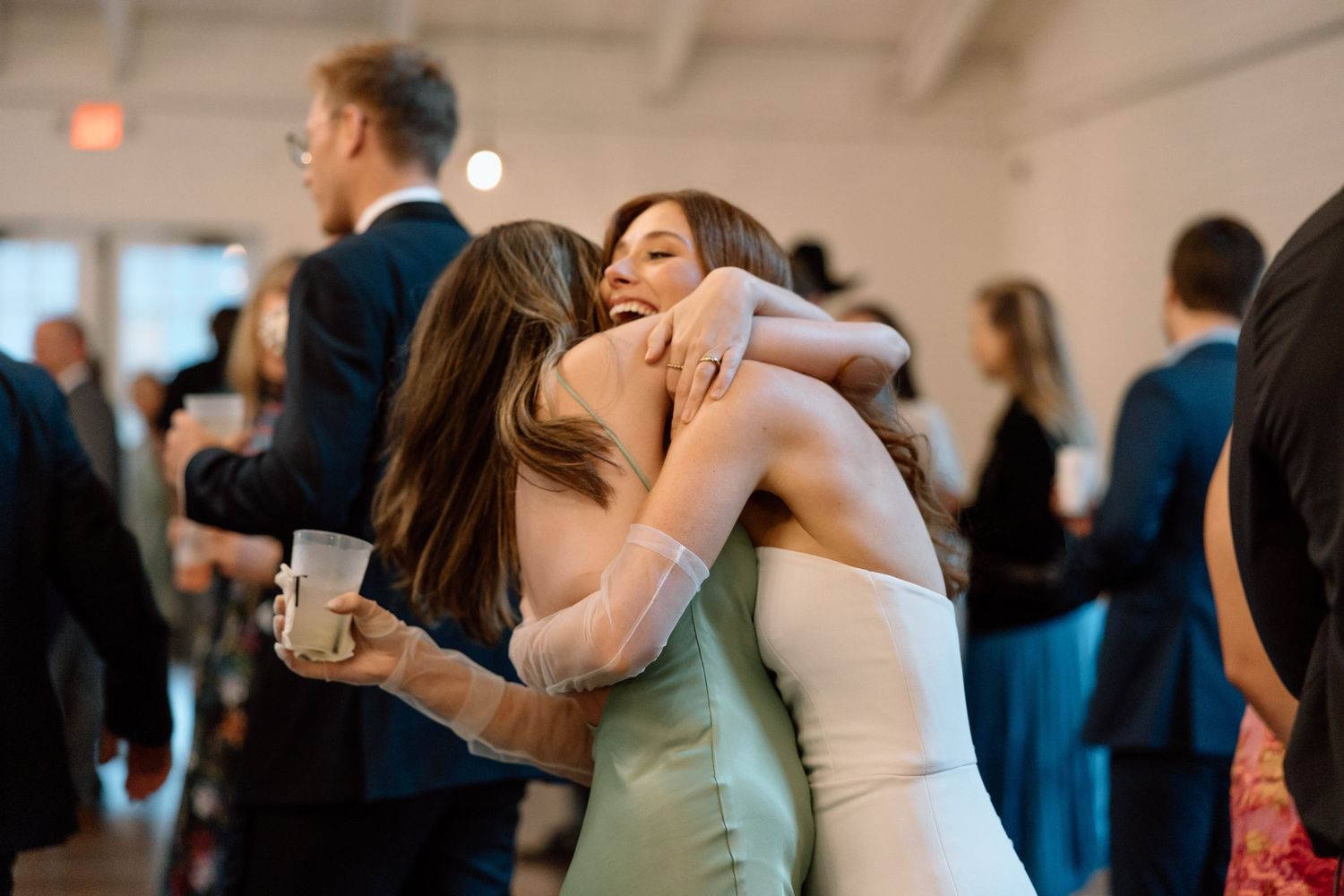 Two people in formal attire share an emotional embrace during a celebration while other guests mingle in the background.