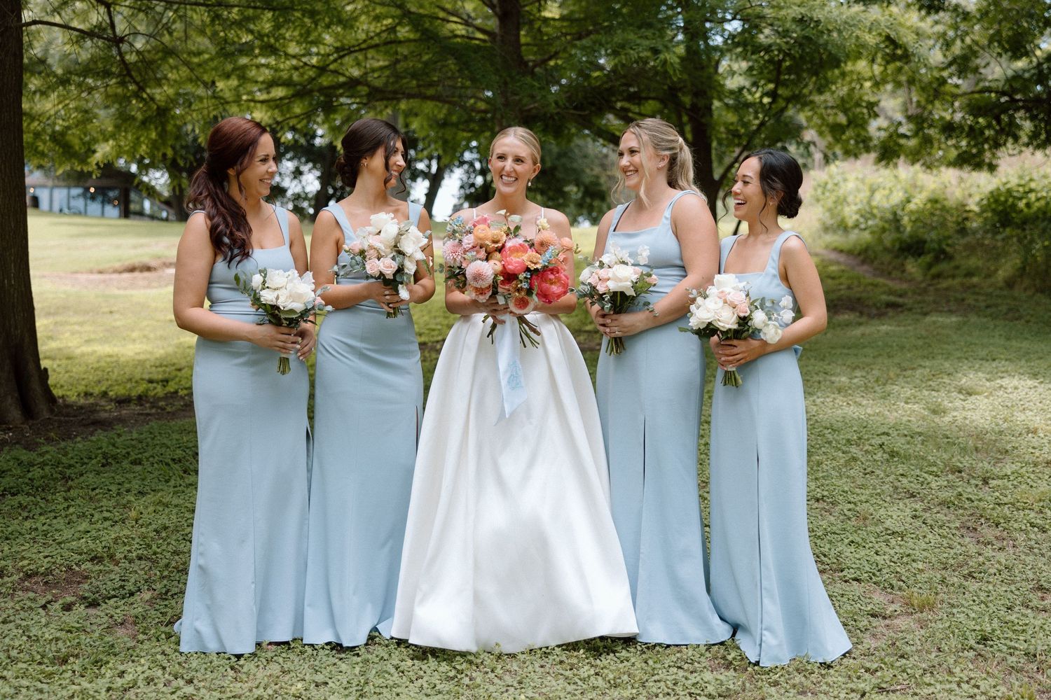 A wedding party in light blue bridesmaid dresses and white bridal gown gather outdoors in a garden setting.