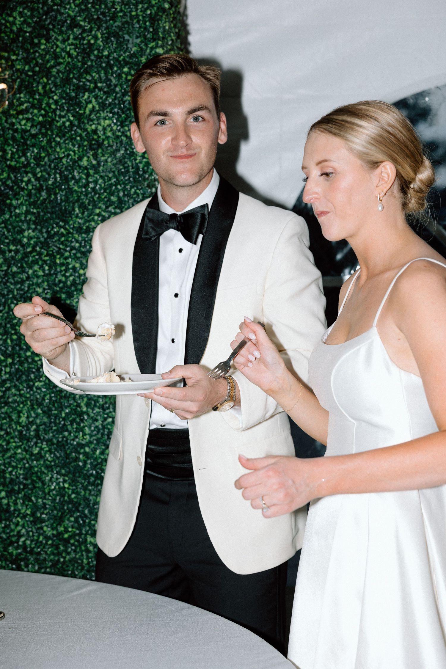 A couple in formal attire, wearing white and black, share cake at an elegant event against a green backdrop.