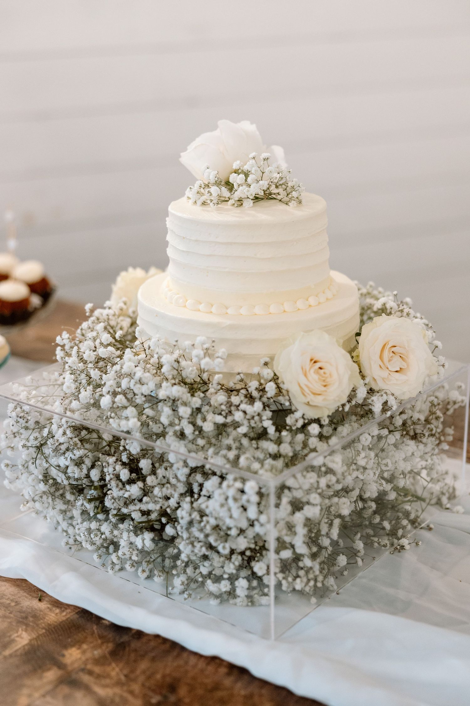 White wedding cake decorated with baby's breath flowers and roses sits on a rustic wooden surface.