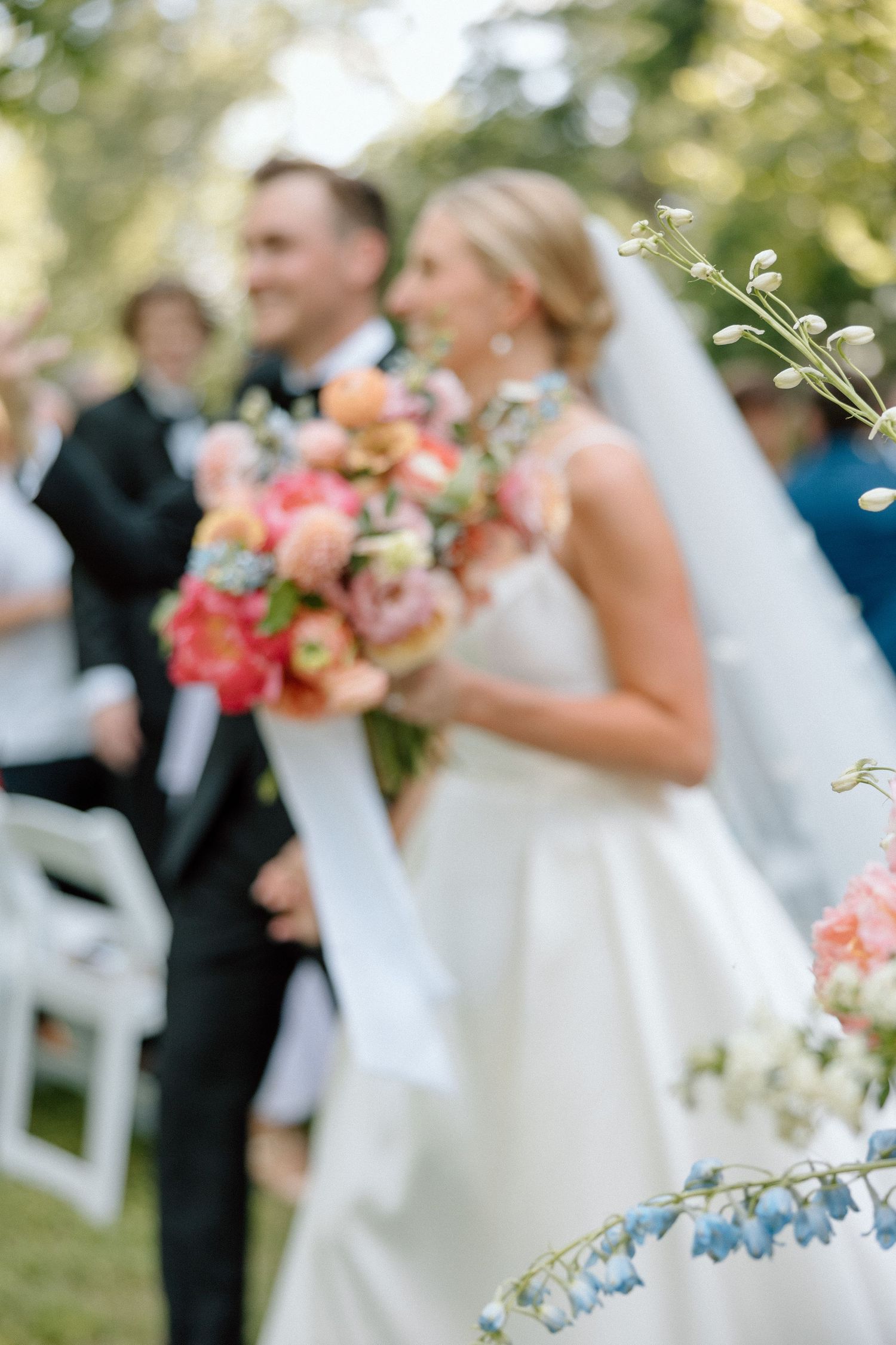 Wedding party stands outdoors with bridesmaids holding vibrant coral and peach bouquets during summer ceremony.
