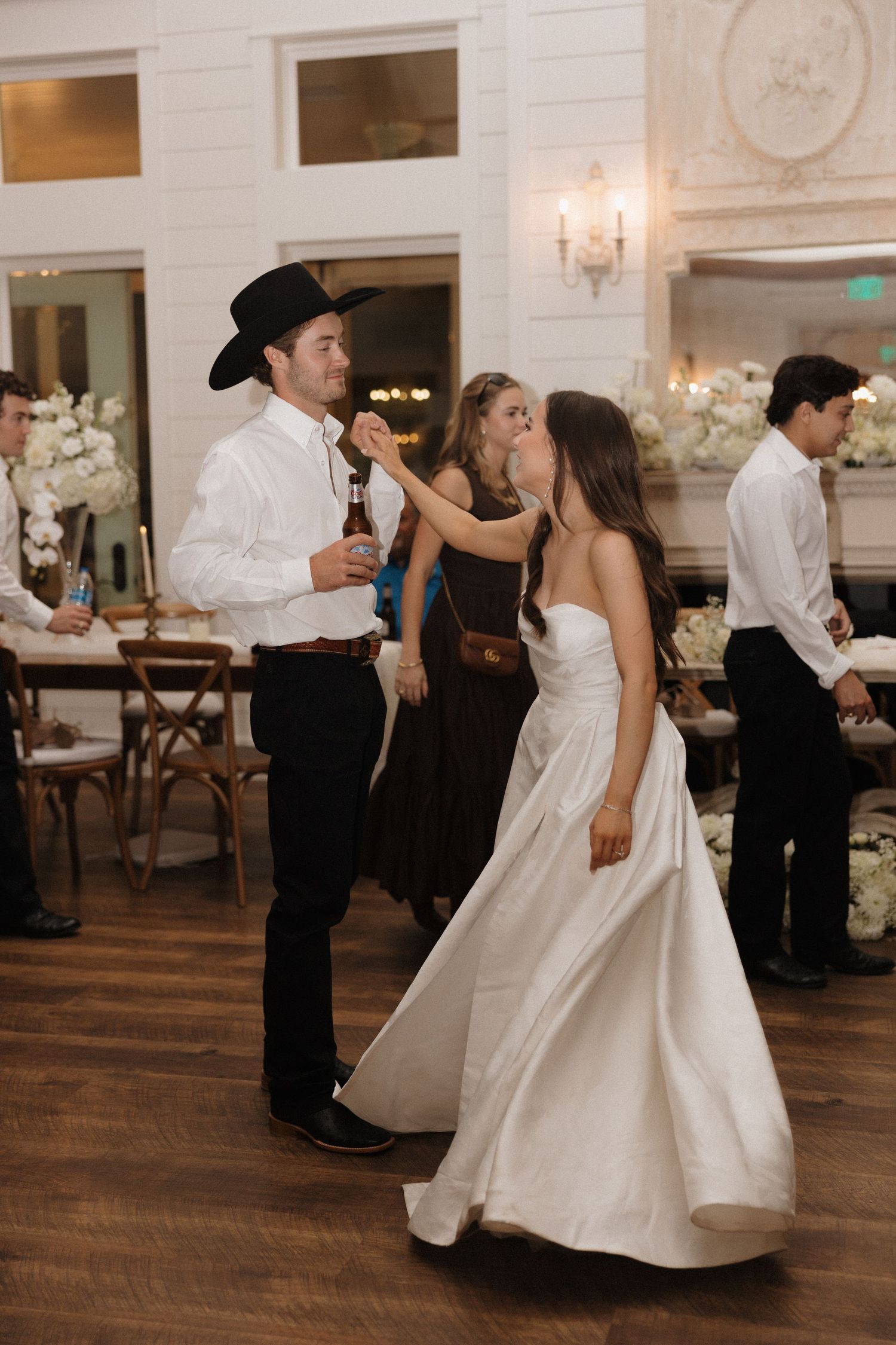 Couple dancing together at an elegant wedding reception with white decor and hardwood floors in a ballroom setting.