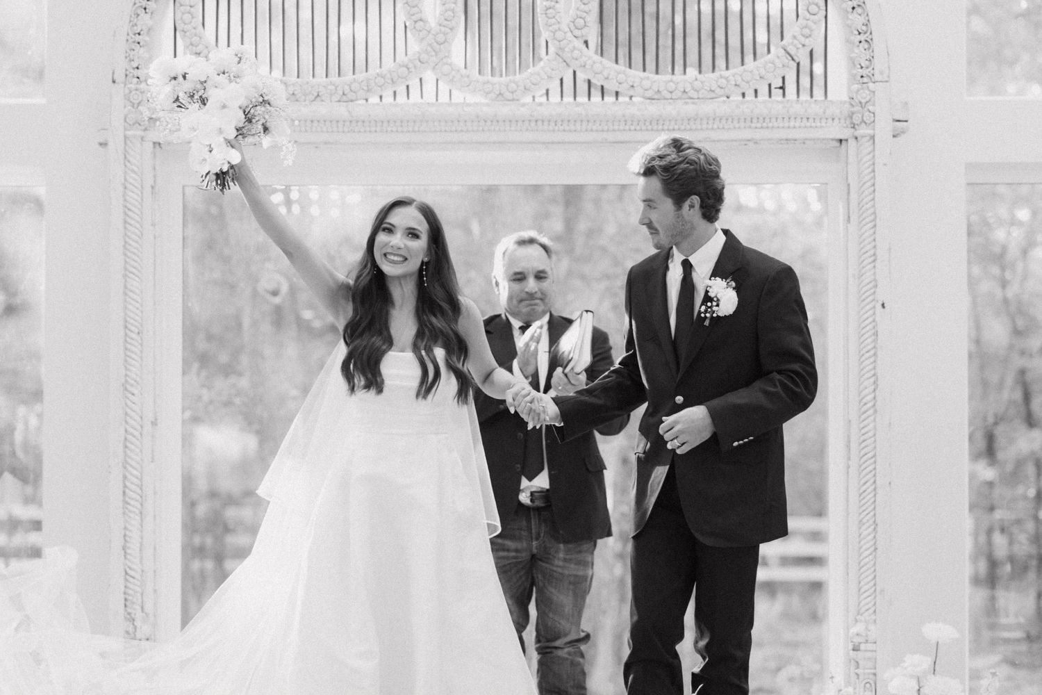 A black and white photo shows a wedding couple walking arm in arm with guests along a stone building exterior.