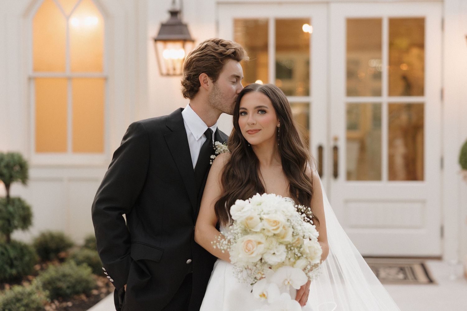 A romantic wedding couple poses outside an elegant white building with glowing lanterns at dusk.