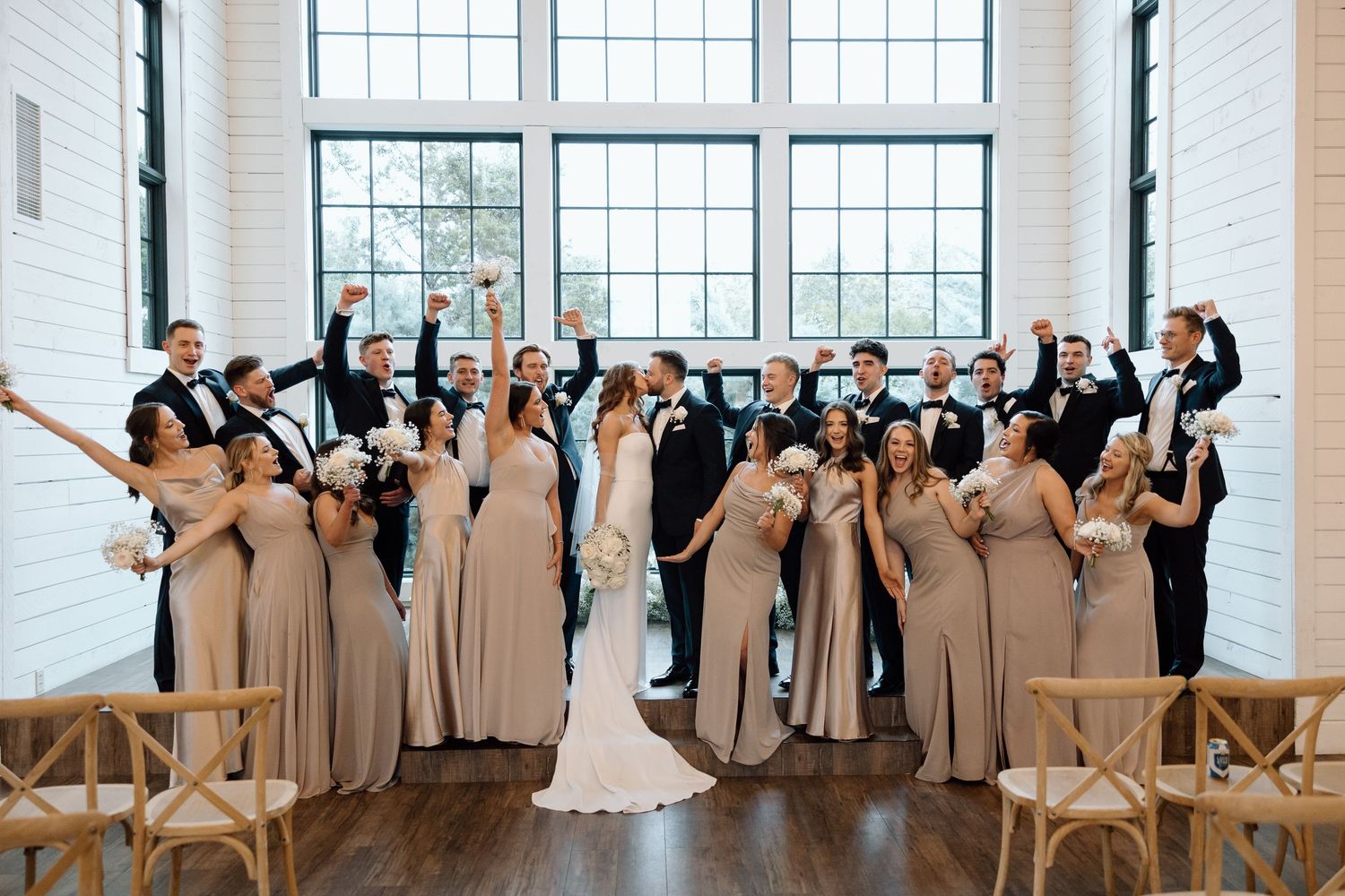 Wedding party poses together in a bright industrial venue with large windows and wooden chairs.