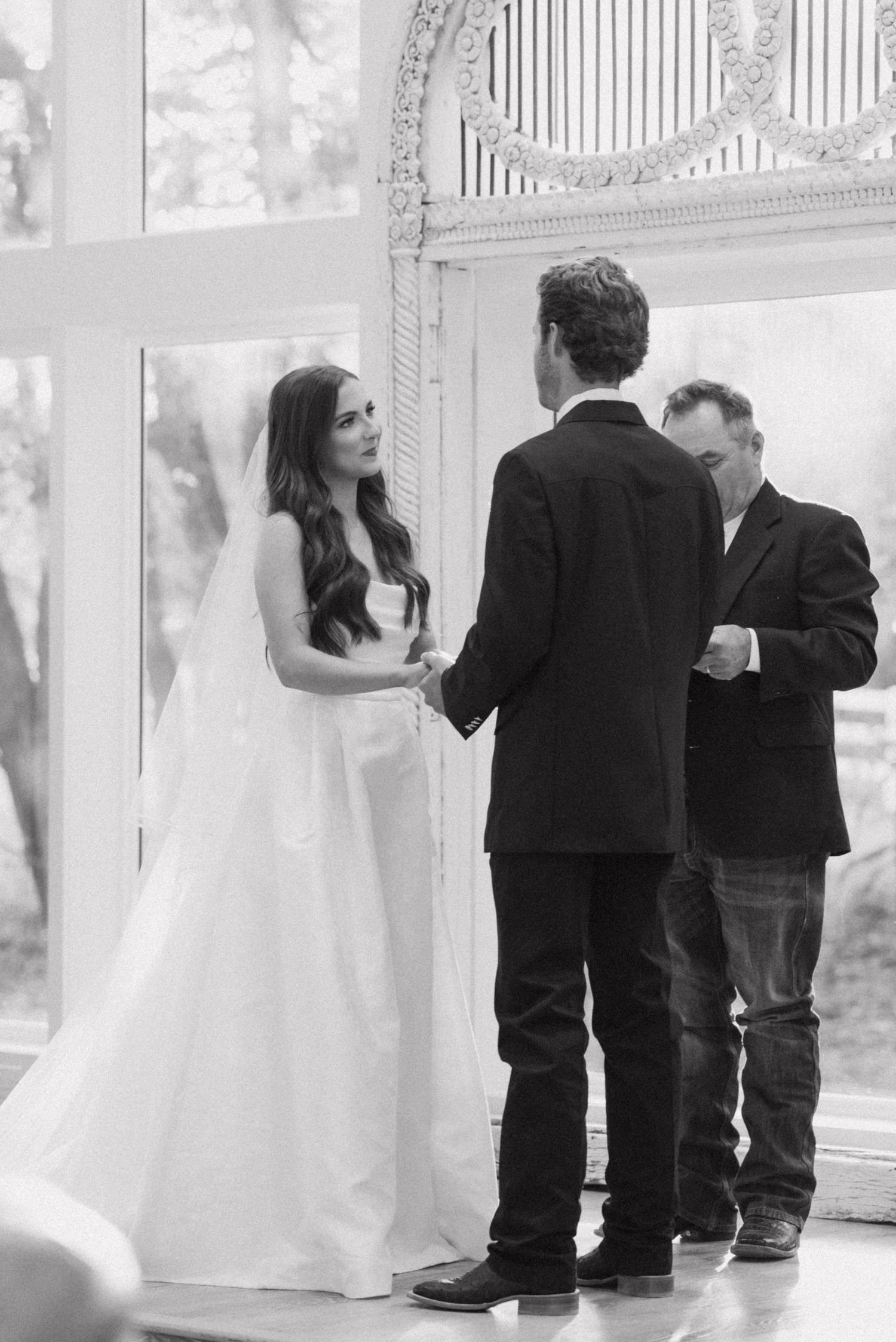 A black and white wedding ceremony photo showing a couple exchanging vows in front of an officiant at an ornate venue.