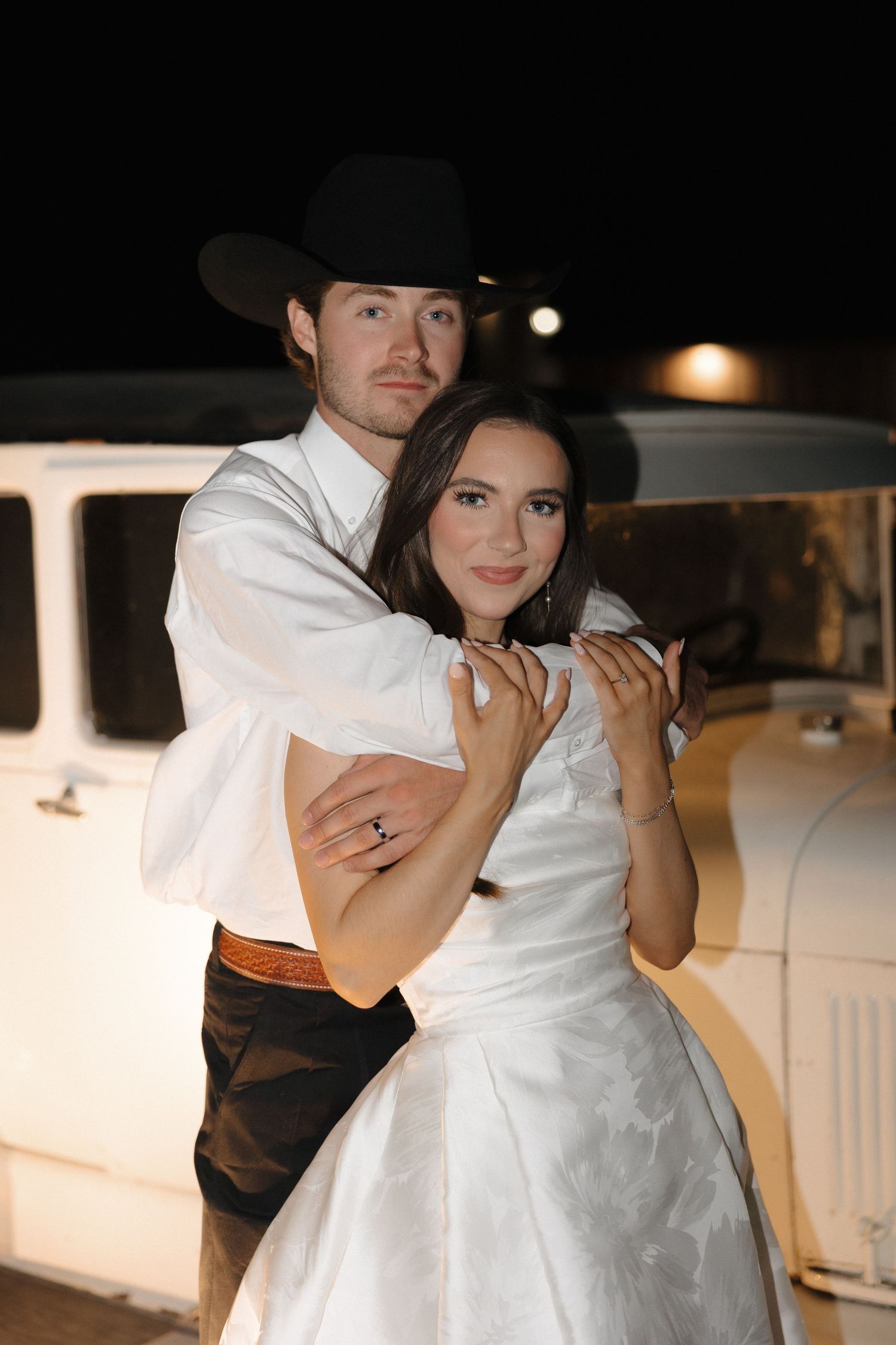 A romantic couple embraces on a dimly lit porch, with the person wearing a white dress and another in dark attire.