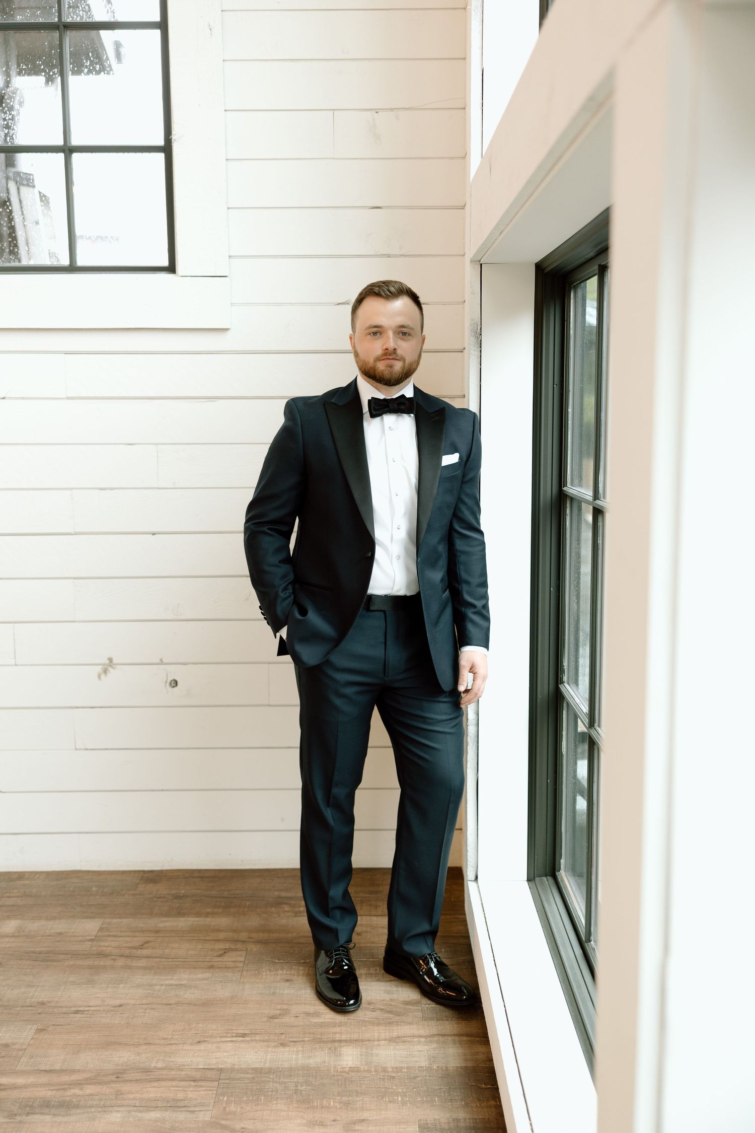 Three views of someone in a classic black tuxedo and bow tie standing in a bright modern hallway with wood floors.