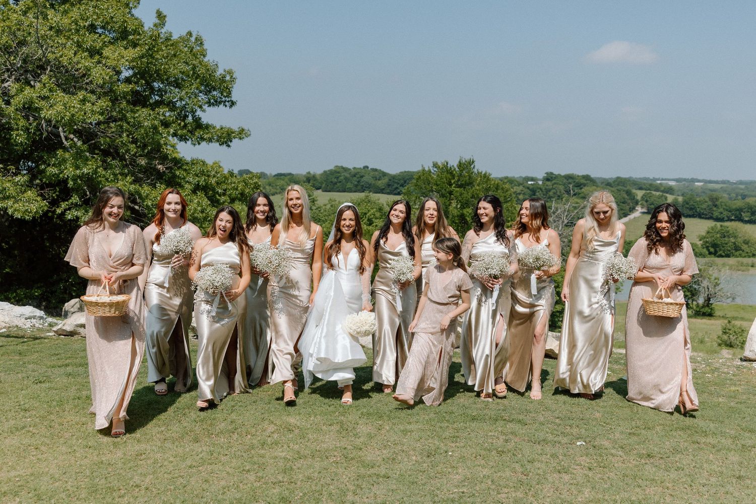 Wedding party in neutral tones poses together on a grassy hillside overlooking scenic countryside views.