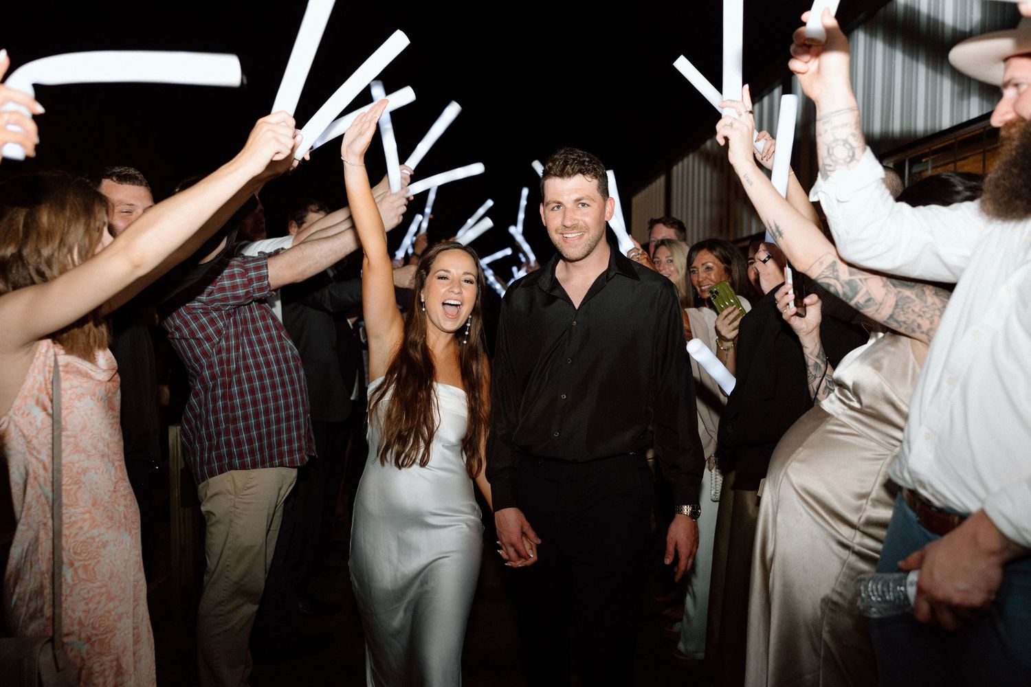 A couple celebrates on a crowded dance floor with guests holding up glowsticks at a wedding reception.