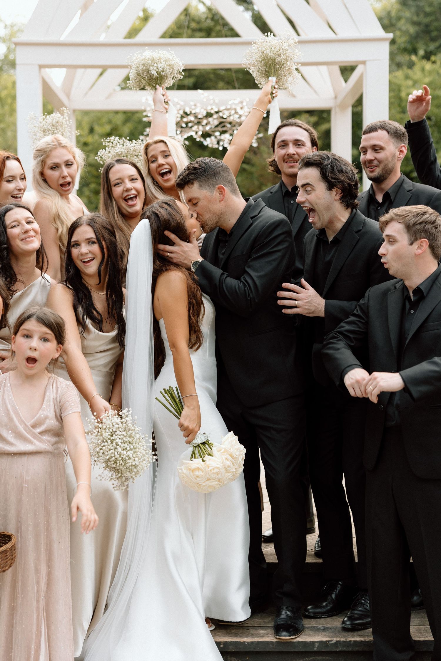 Wedding party celebrates joyfully together under white arch decorated with baby's breath flowers during outdoor ceremony.