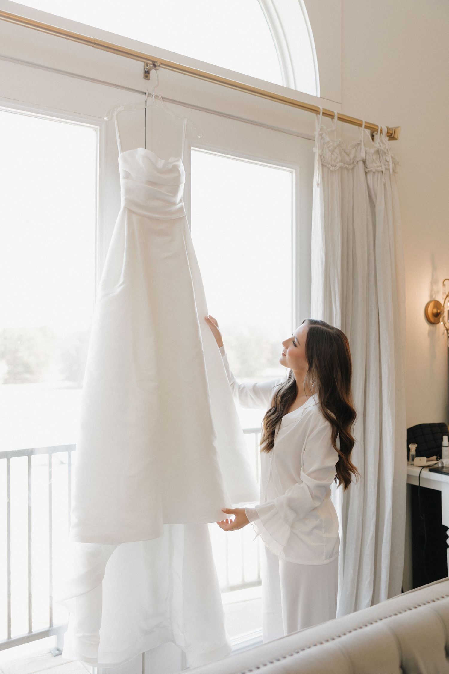 A person in white clothing hangs sheer white curtains on tall windows in a bright, sunlit room.