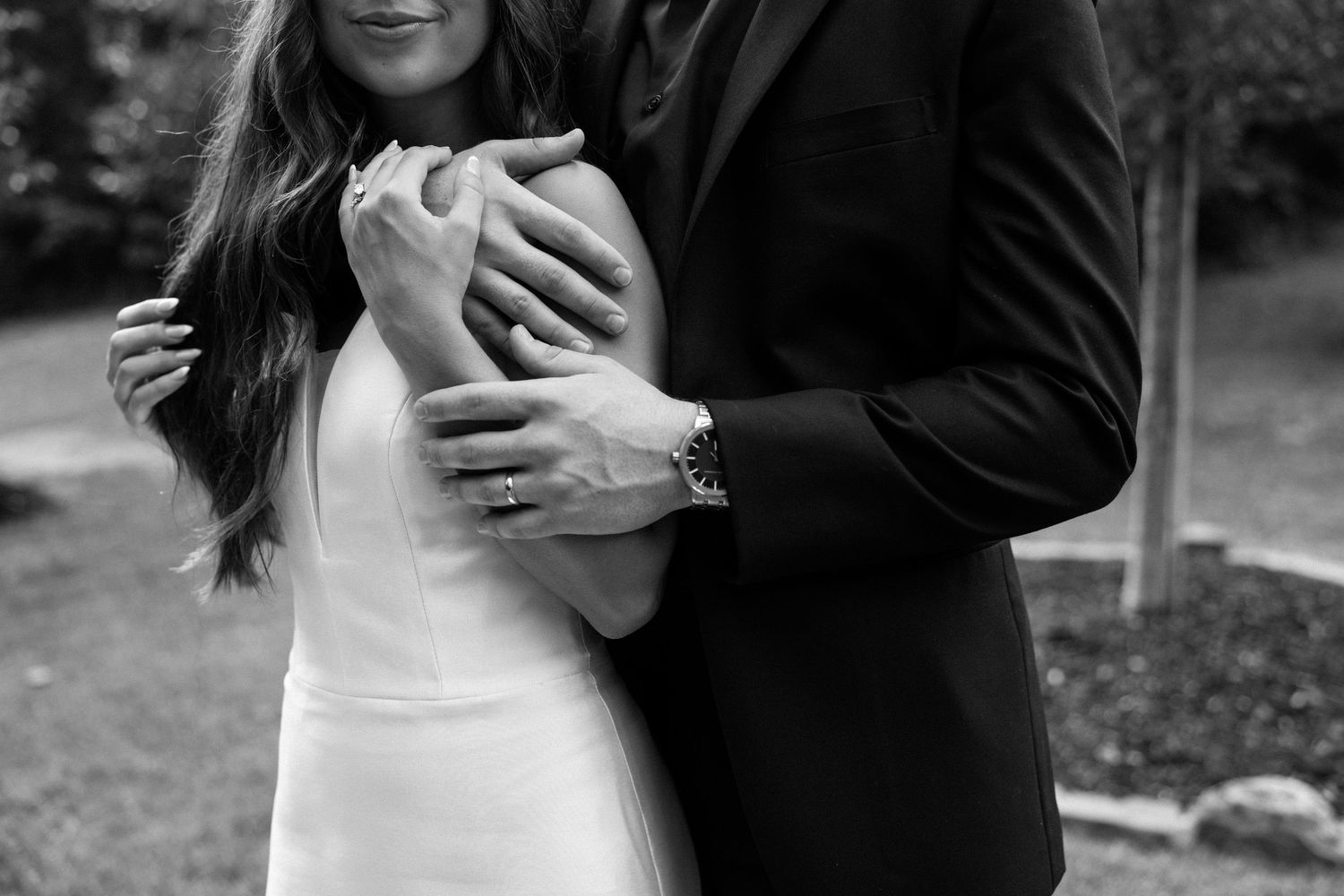 A romantic couple in formal wedding attire embraces outdoors in a garden setting.
