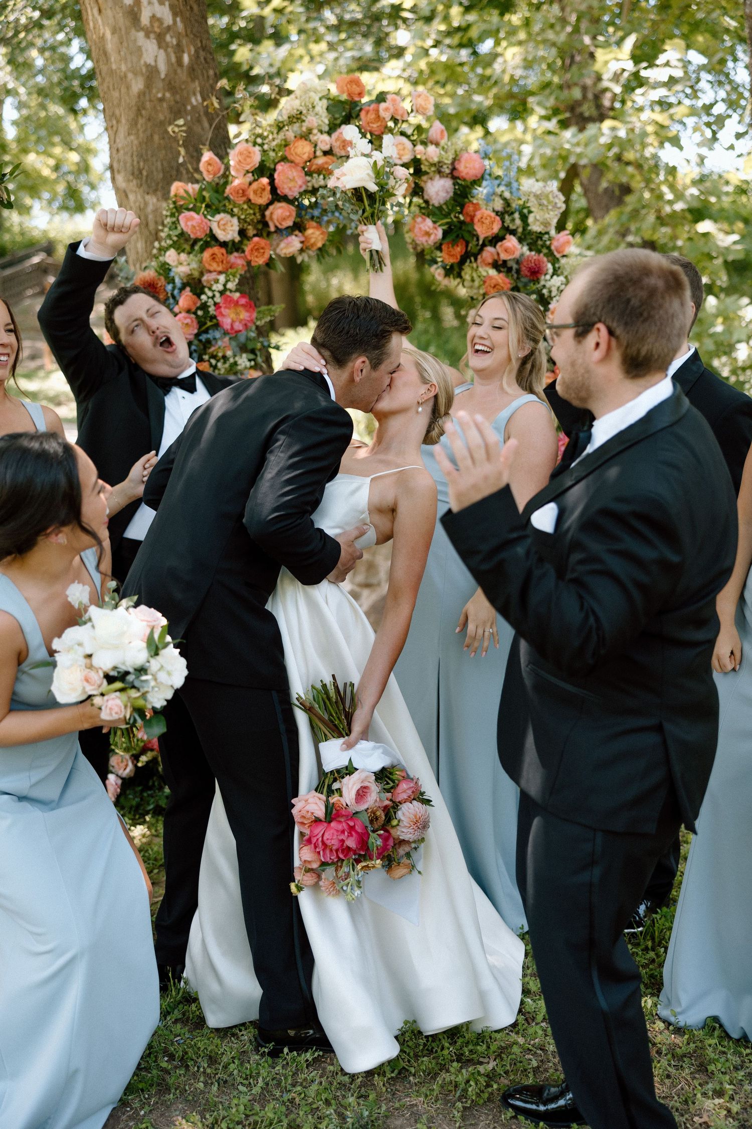 Wedding party in formal attire poses together outdoors with bridesmaids in light blue dresses and groomsmen in black suits.