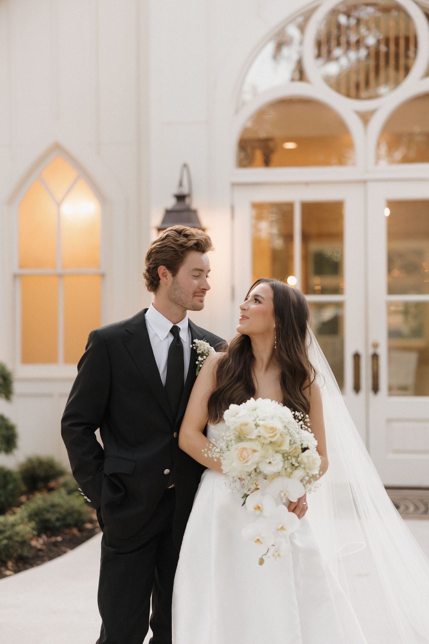 A bride and groom share romantic moments in front of white arched windows at an elegant wedding venue.