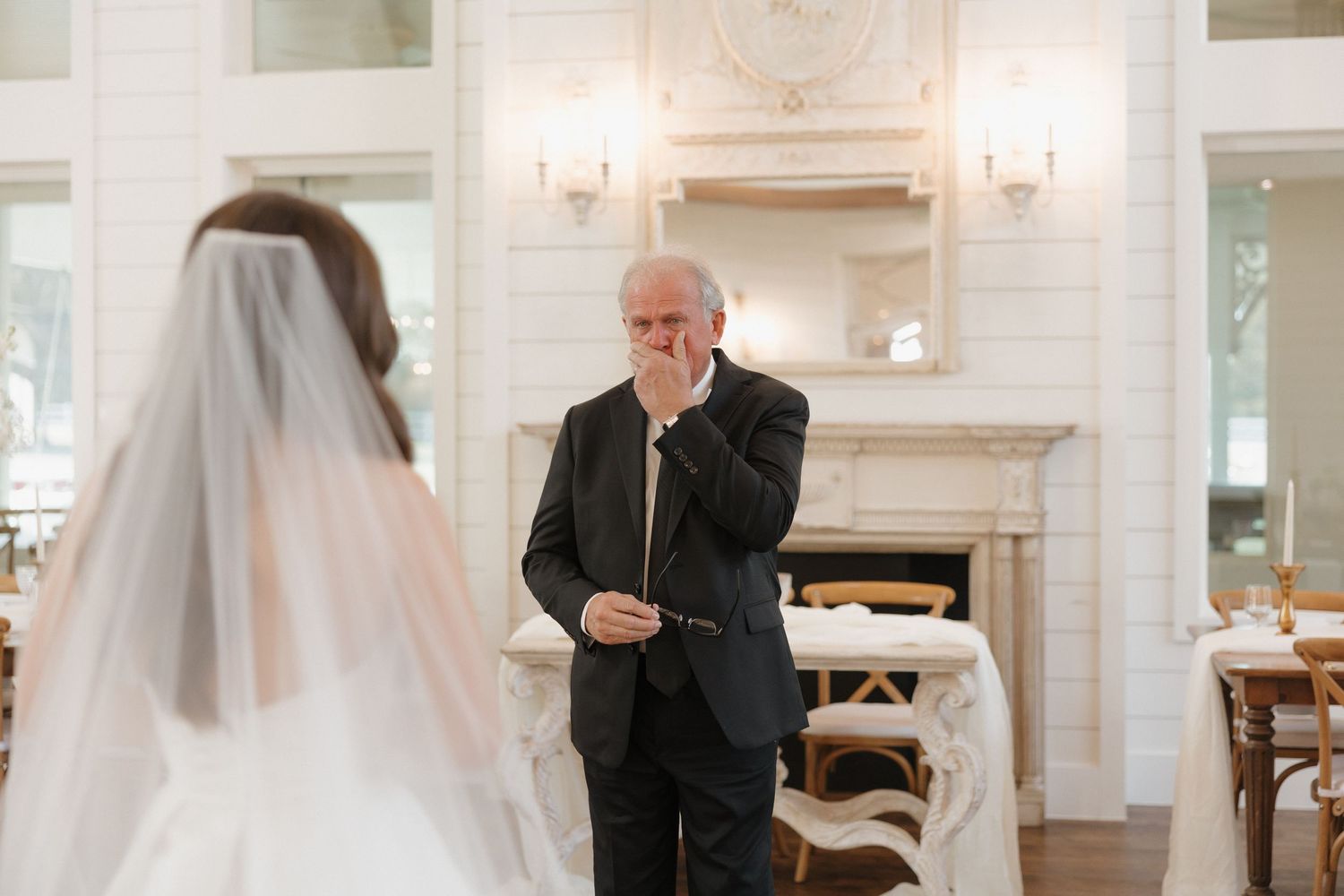 A couple in formal wedding attire shares an emotional moment in an elegant white room with chandeliers.
