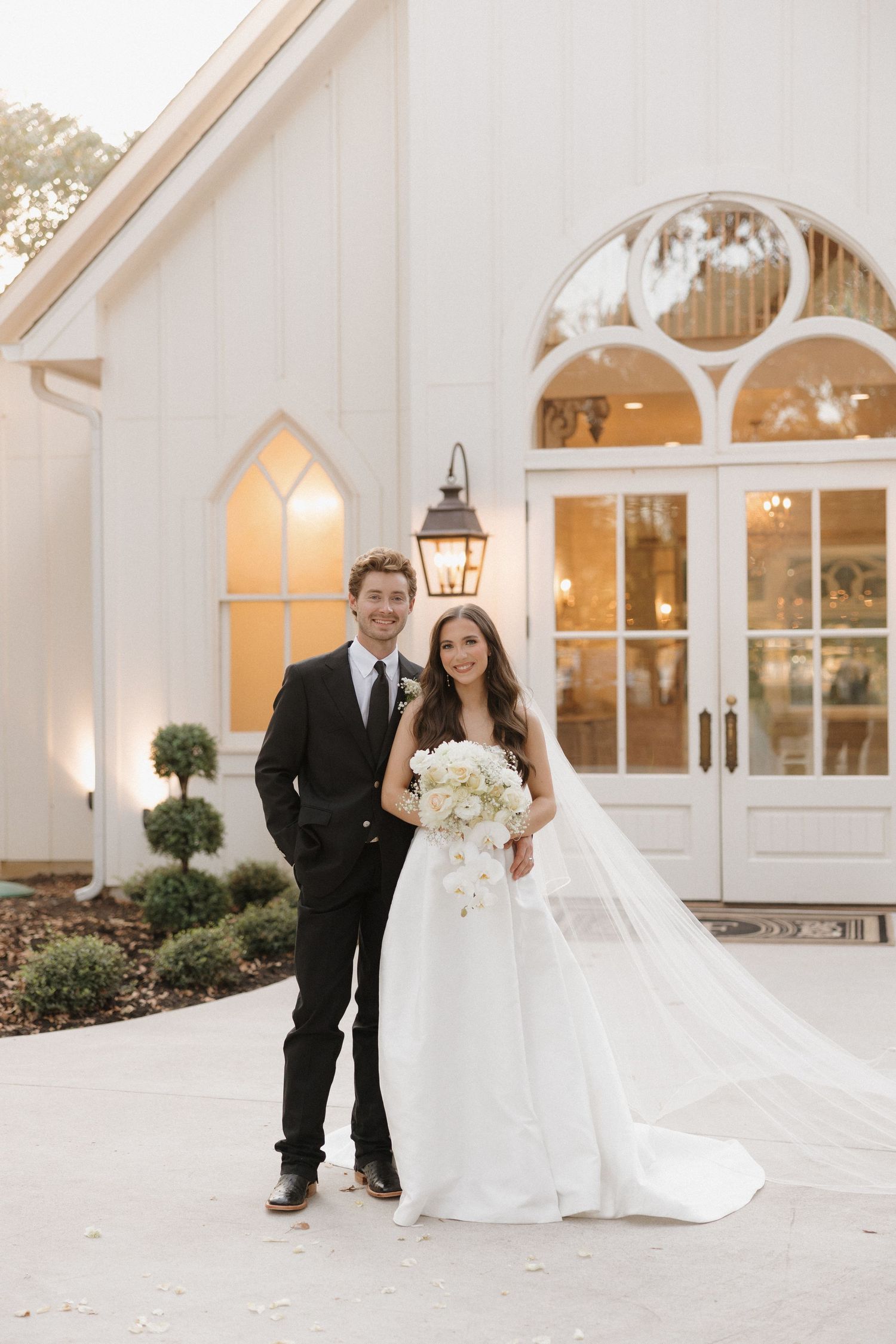 A couple stands outside a white chapel with arched windows and elegant French doors on their wedding day.