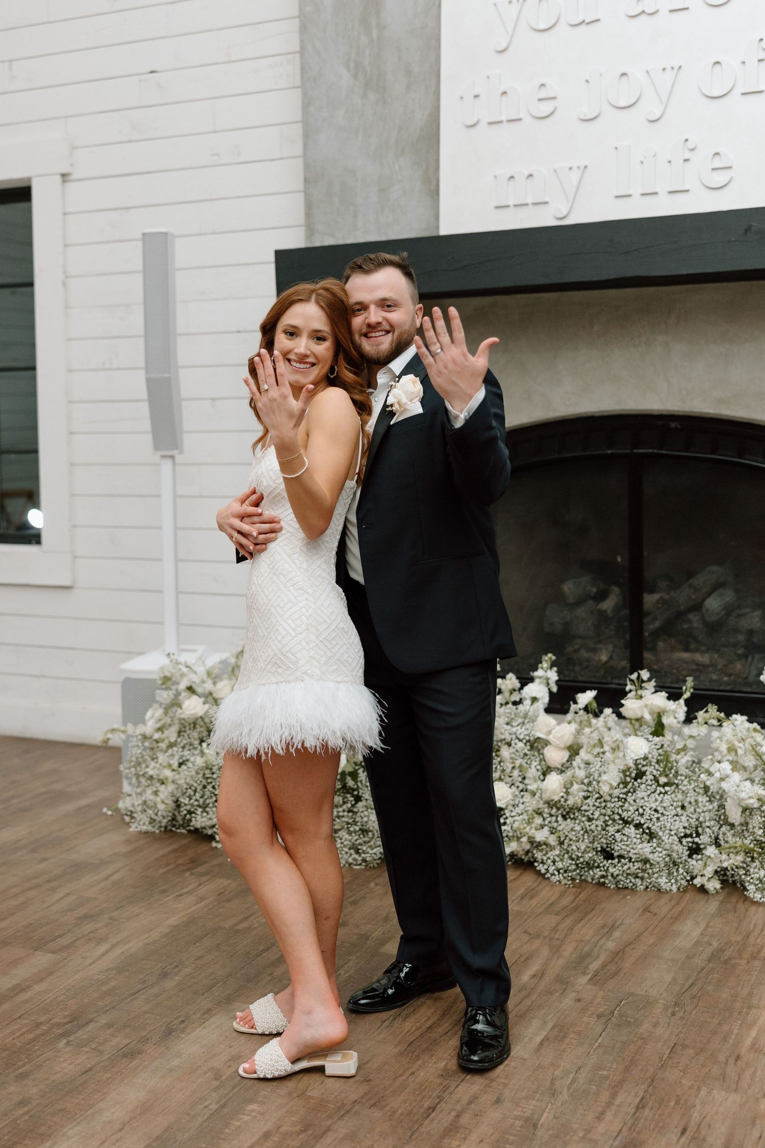A couple celebrates at their wedding venue, with the bride in a feathered white mini dress and groom in classic black suit.
