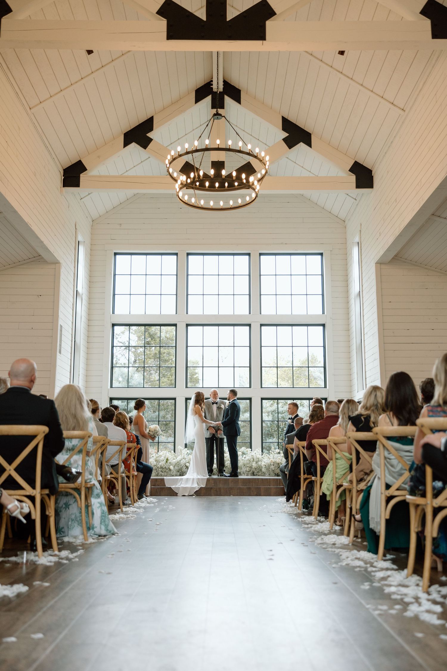 Interior of a modern white chapel with vaulted ceiling, large windows and rustic chandelier during a wedding ceremony.