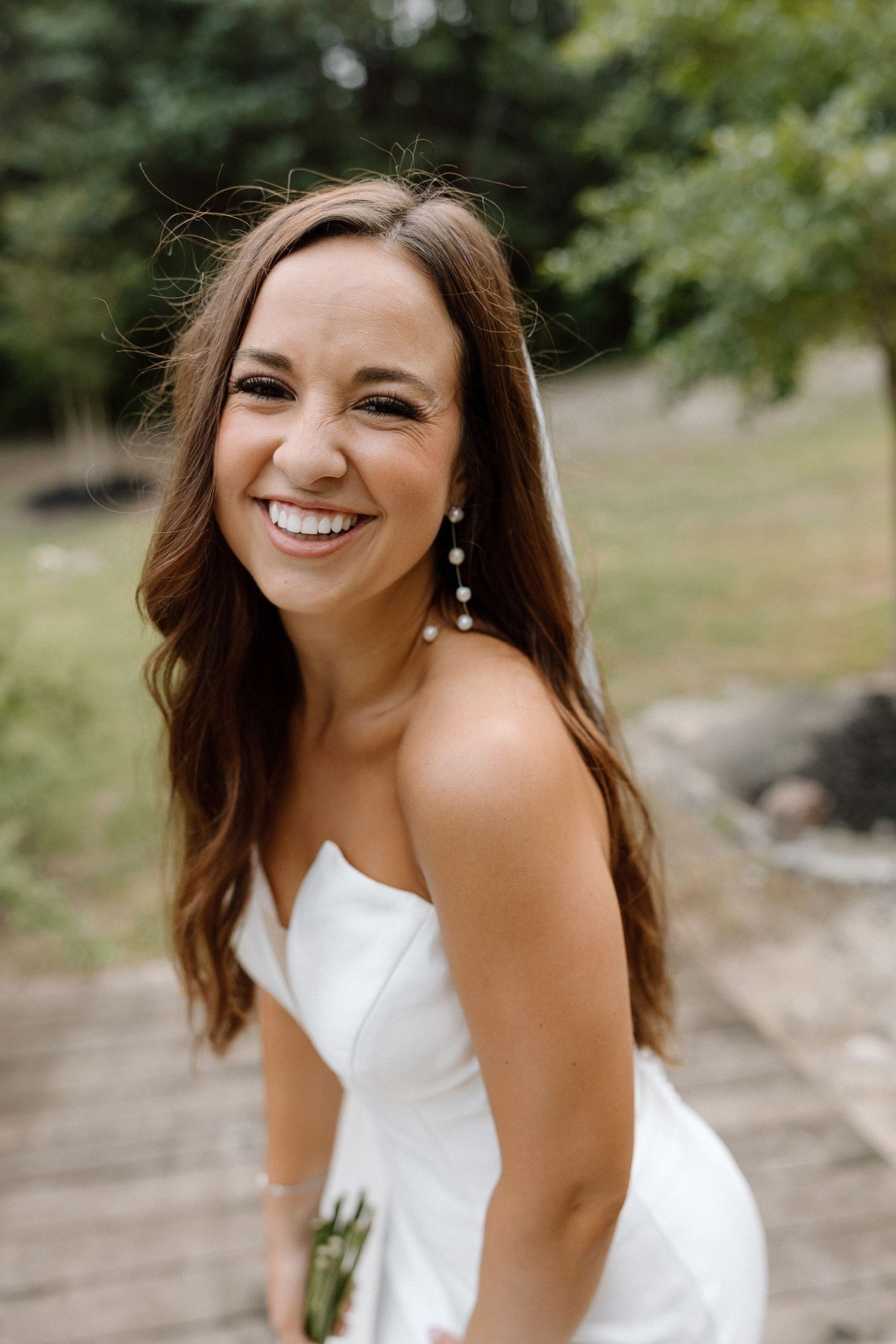 A person wearing a white sleeveless dress smiles joyfully in an outdoor setting with trees in the background.