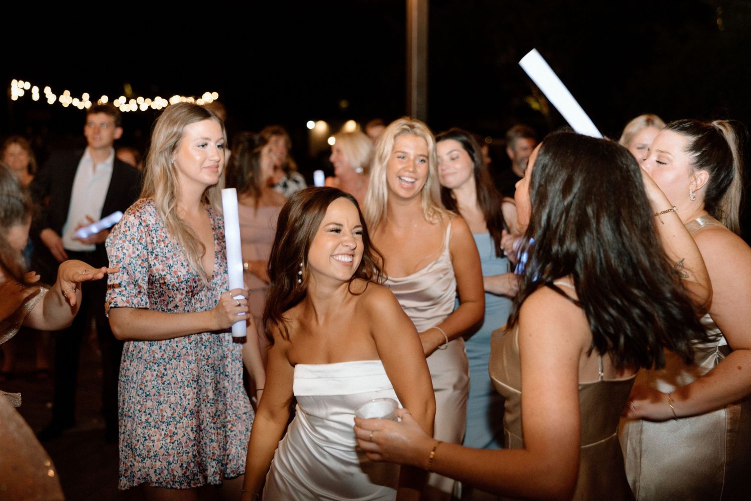 Party guests dancing and celebrating at an outdoor evening event with string lights overhead.