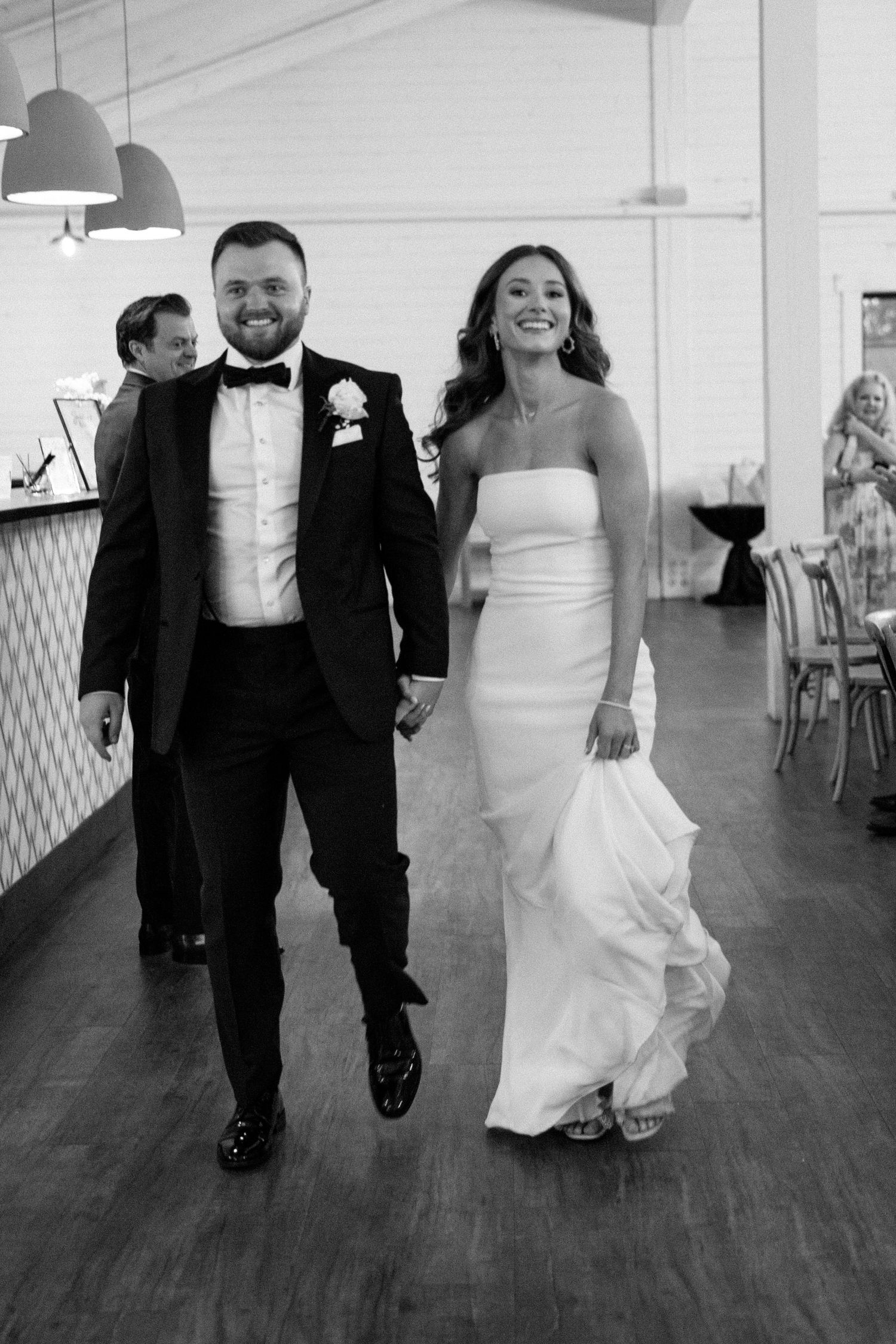 A black and white wedding photo of a couple walking together indoors, the bride in white and groom in tuxedo.