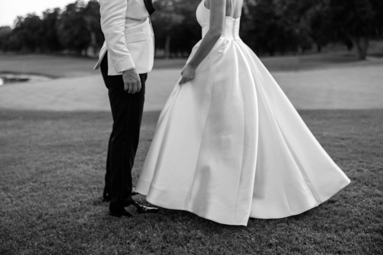Black and white photo showing bottom half of couple on grass, with groom in dark pants and bride in flowing white gown.