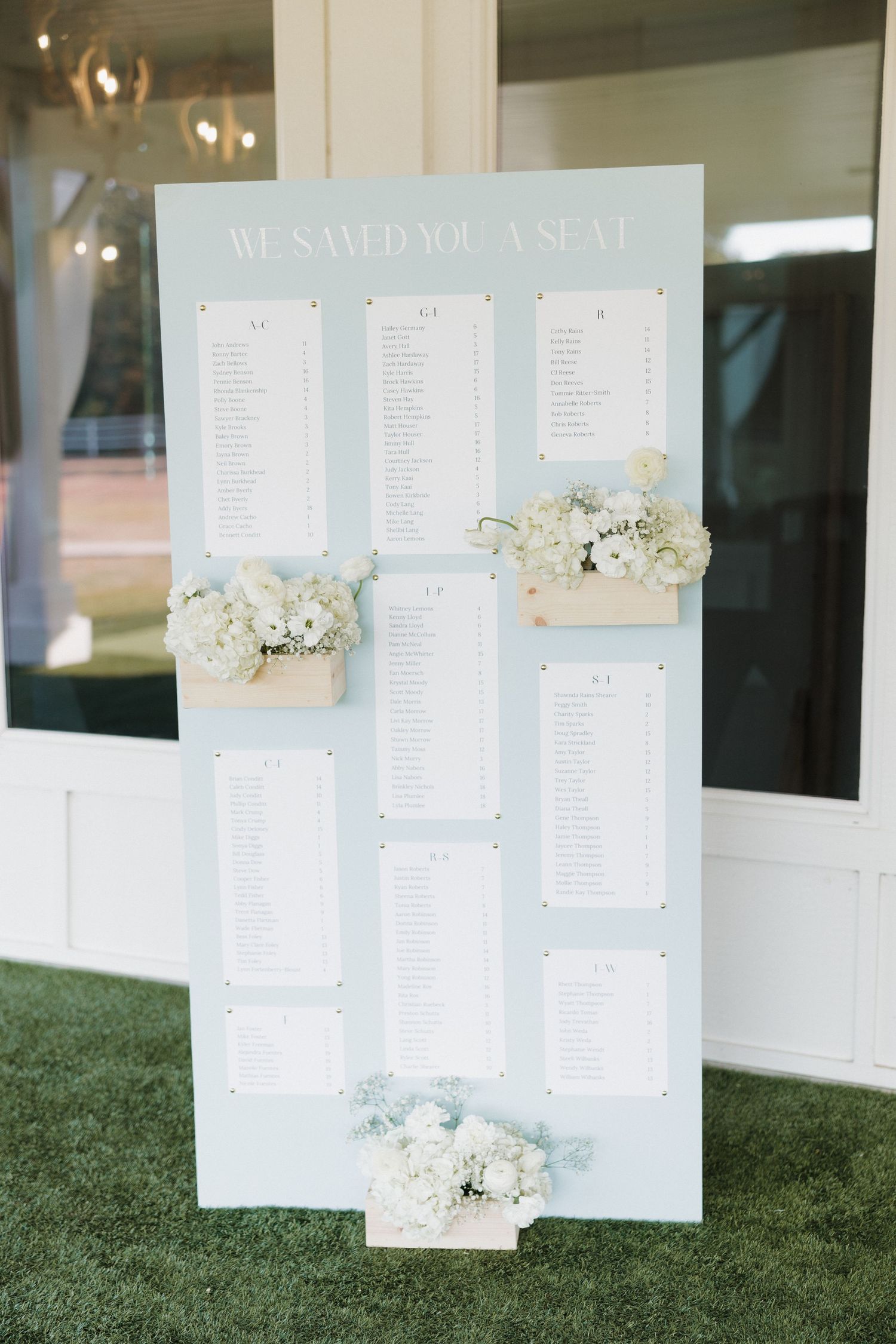 Wedding seating chart display with white paper lists mounted on grass with delicate floral arrangements.