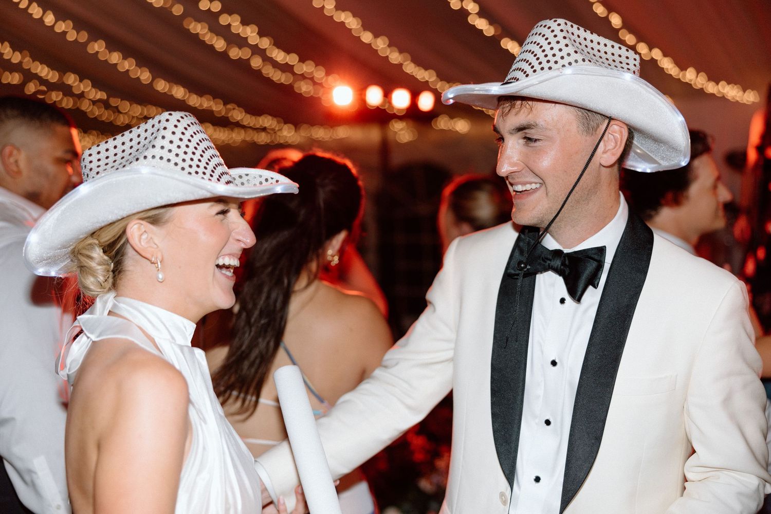 People in white cowboy hats and formal attire dance together at an event with string lights overhead.
