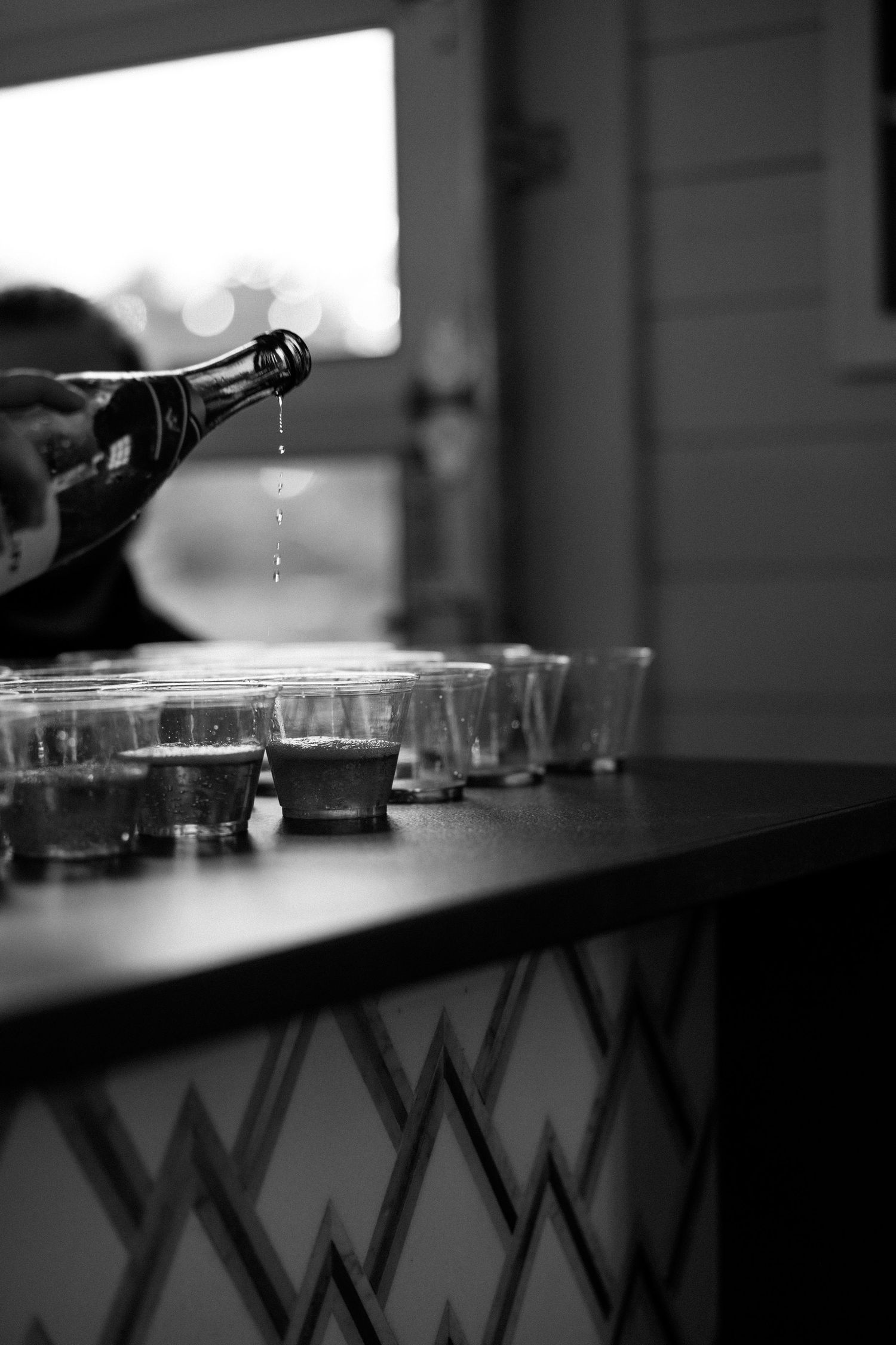 Black and white close-up of glasses and bottles on a bar counter in dim lighting.