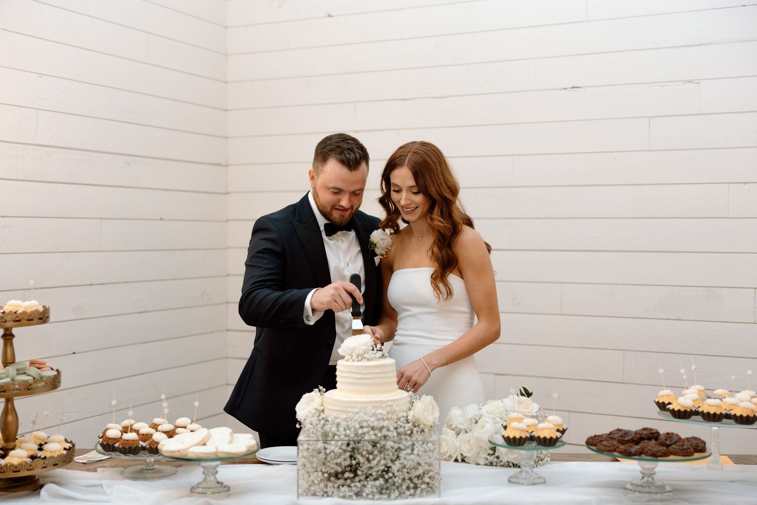 A wedding dessert table features an elegant white cake alongside cupcakes and cookies in a bright white room.