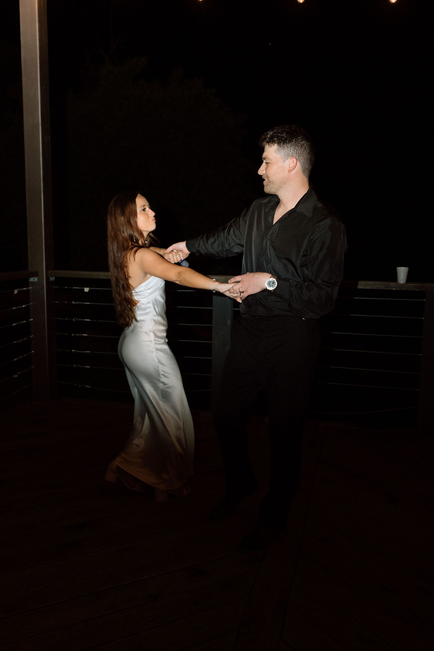 A couple dances together at night on a dark balcony, the woman in white and the man in black sharing an intimate moment.