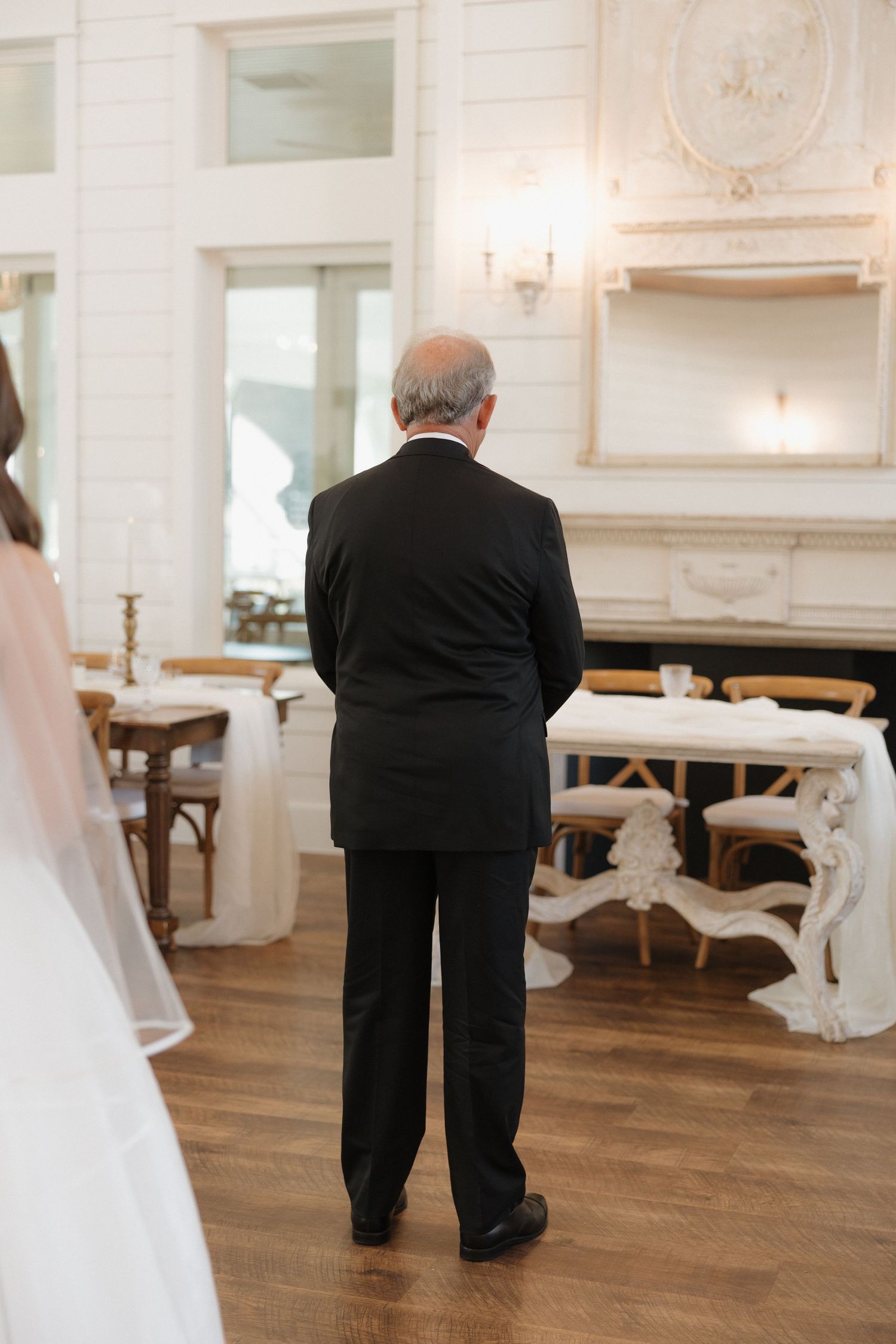 A person in a black suit stands alone in an elegant white-walled room with hardwood floors and ornate decor.