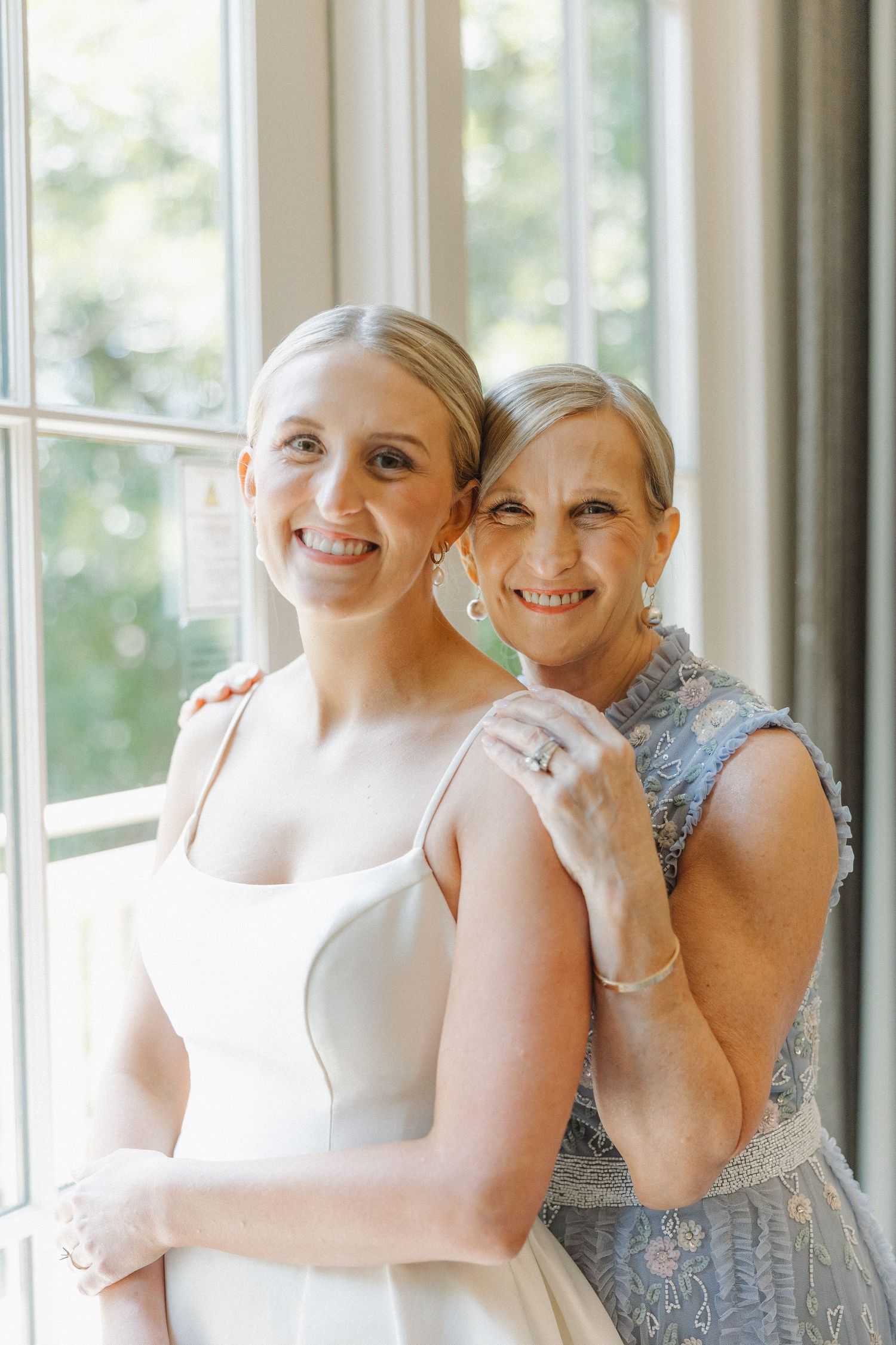 Two women in formal attire share a tender moment by a sunlit window, wearing a white gown and blue lace dress.
