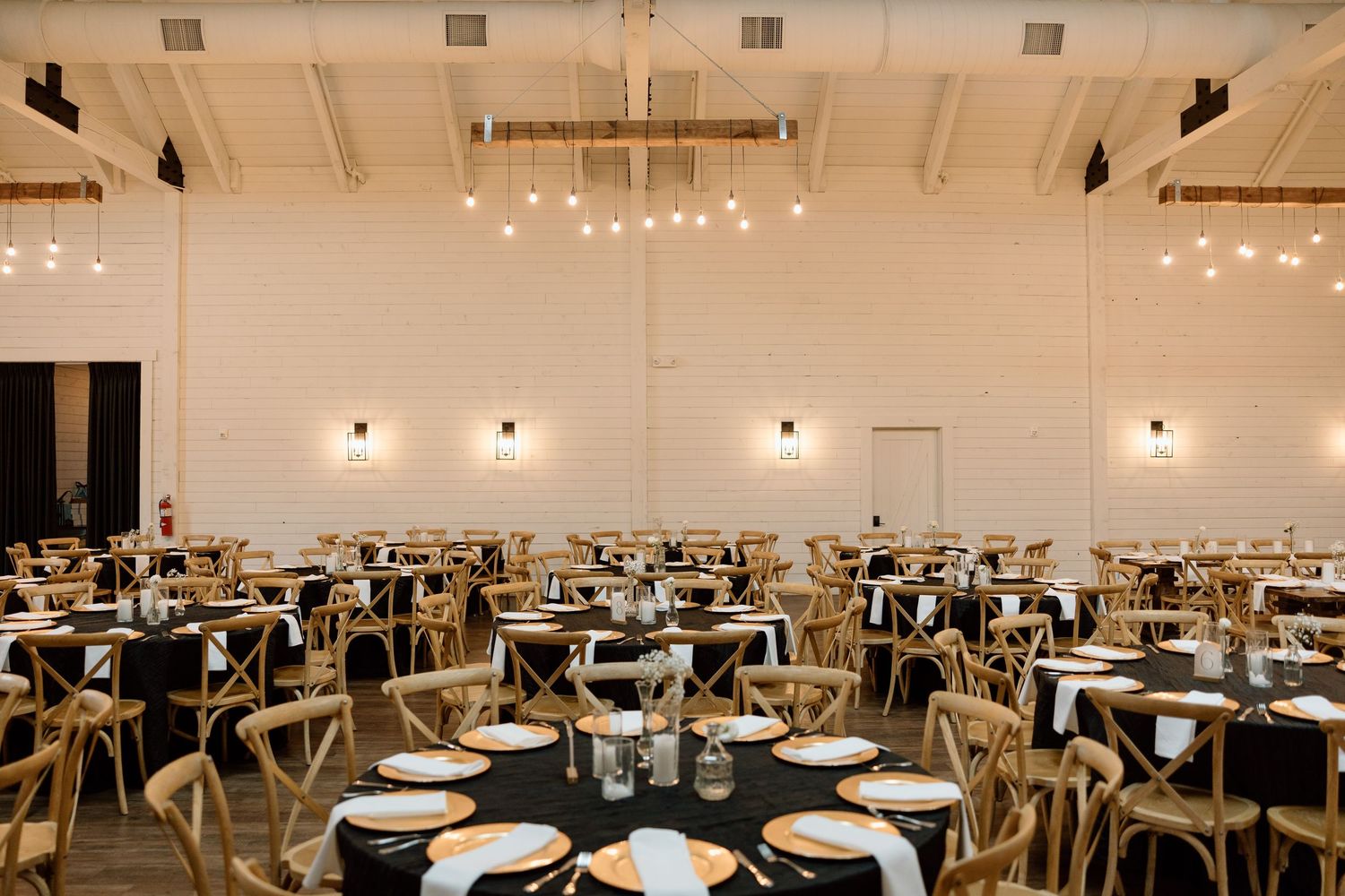 Modern event venue with round tables and wooden chairs set for dinner under industrial ceiling with string lights and sconces.