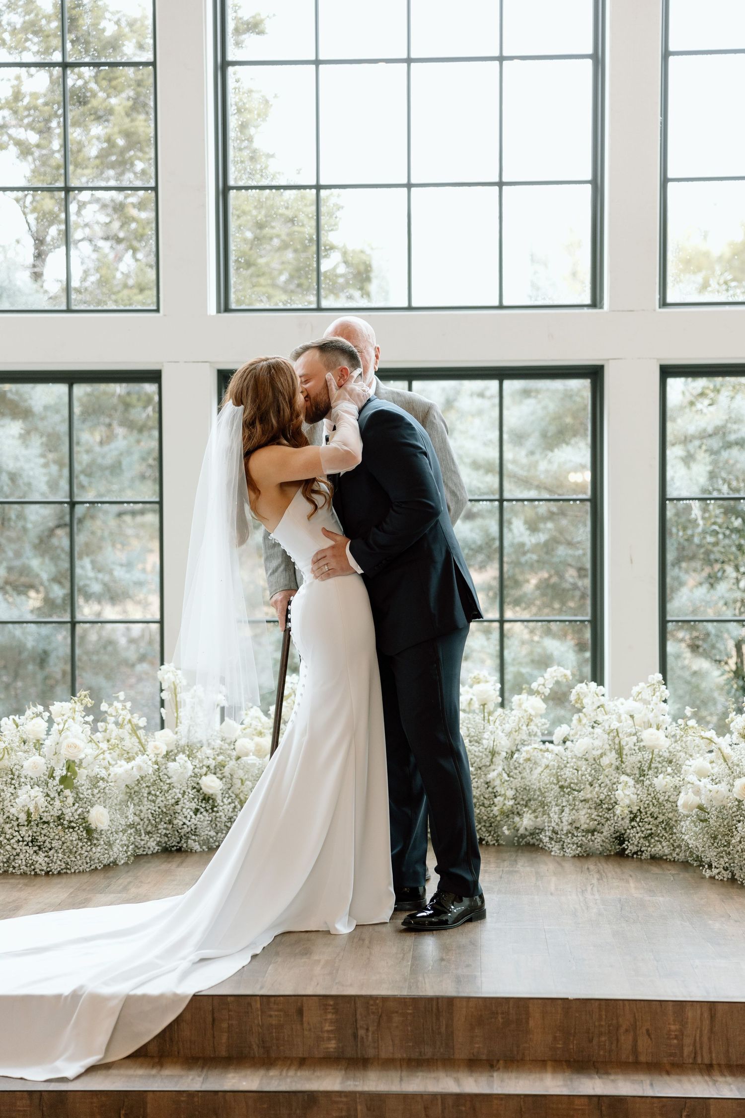 Romantic wedding kiss in front of large windows with white floral arrangements decorating the venue.