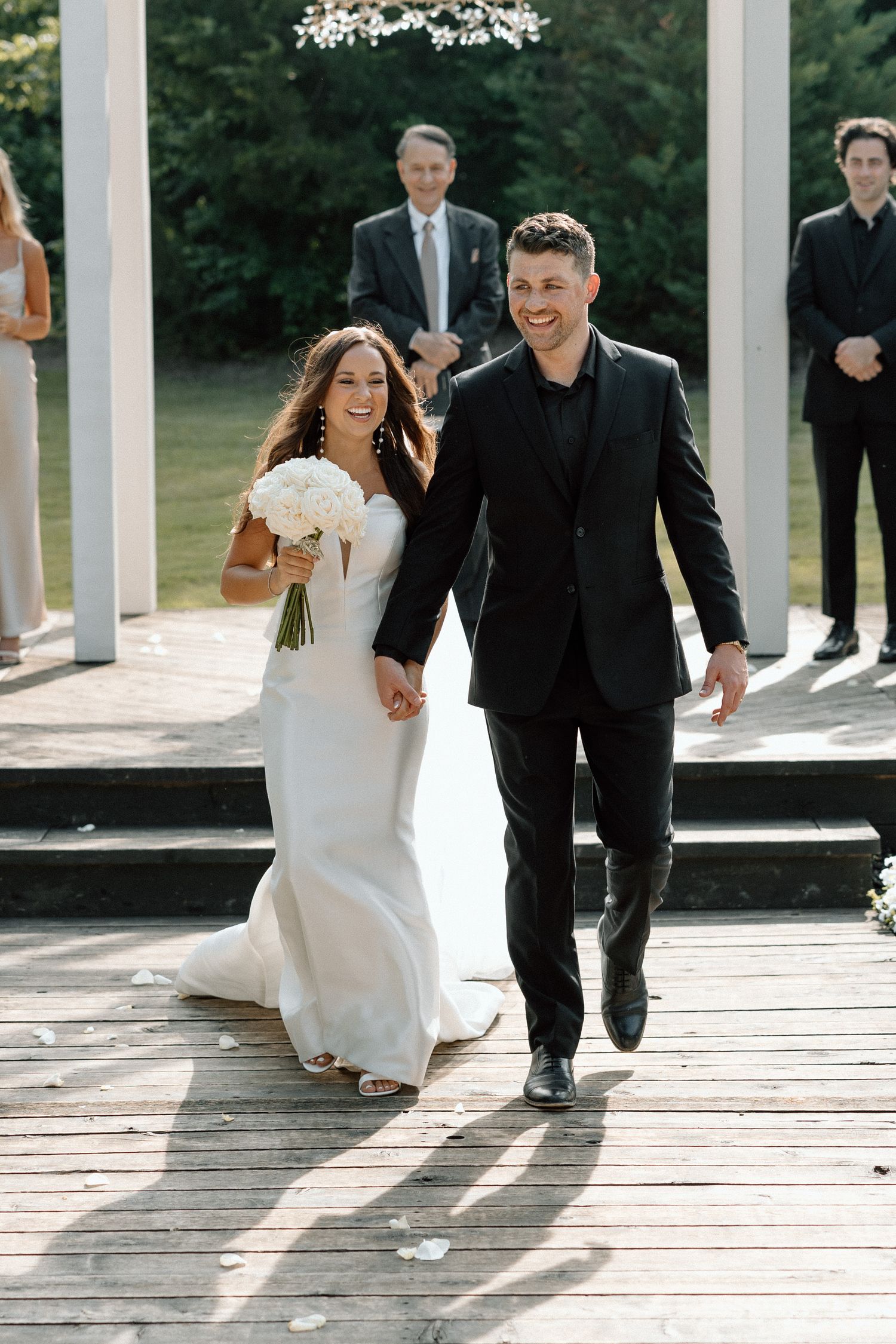 A couple walks together on a wooden deck surrounded by white pillars on a sunny summer day.