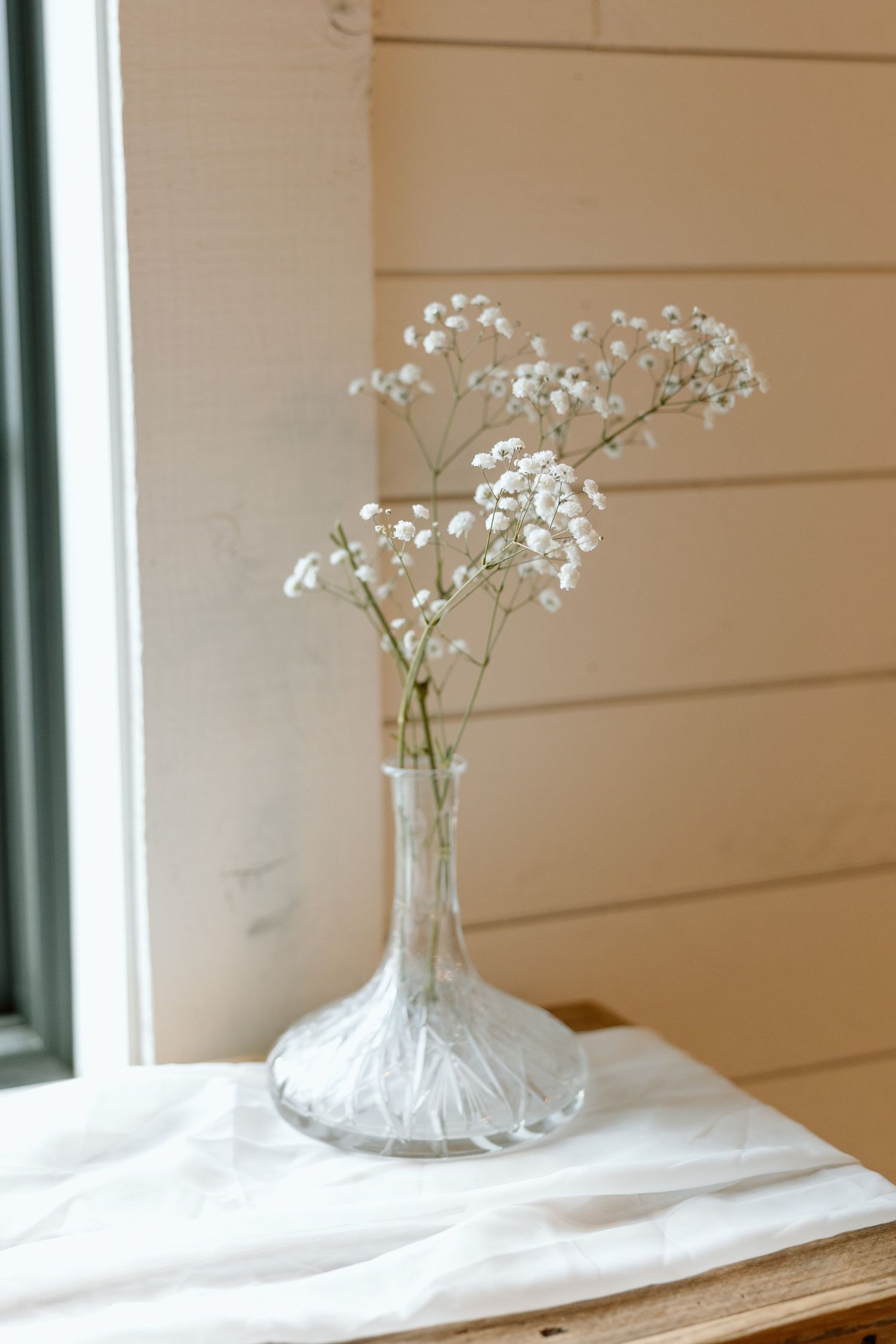 Delicate white baby's breath flowers arranged in a clear glass vase with a textured base on a white cloth by a window.