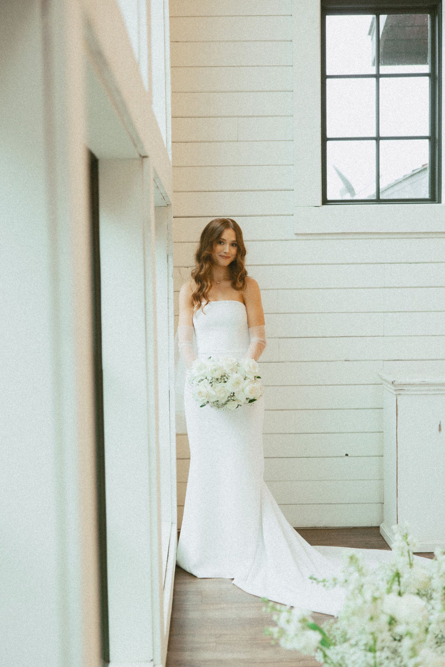 A bride in a white strapless wedding gown poses gracefully in a bright, white-walled room with natural light.