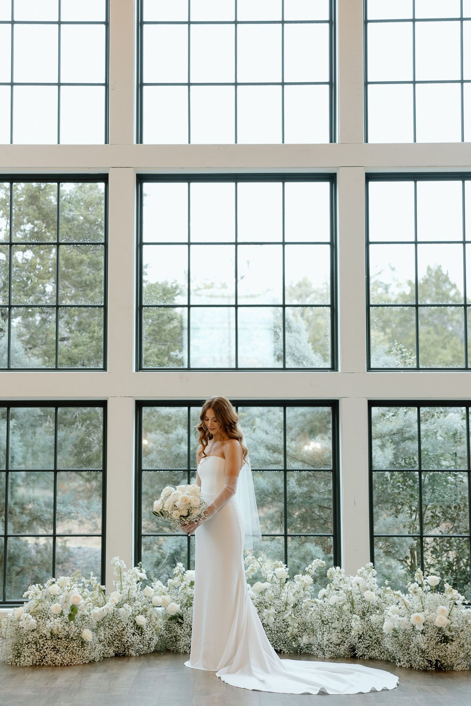 A bride in a white gown stands in front of floor-to-ceiling windows with a floral installation in a modern venue.