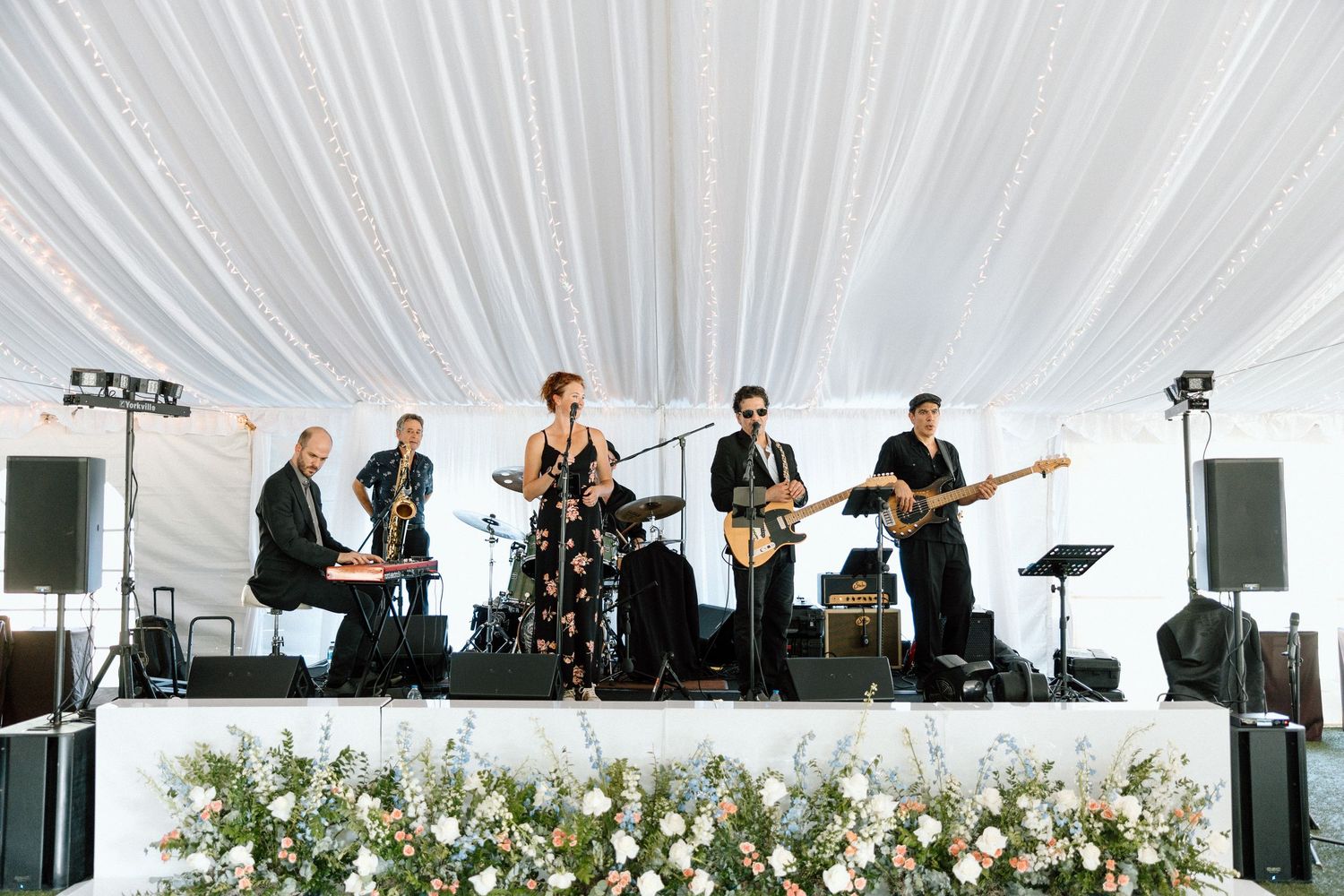 Live band performs on stage under a white tent at an outdoor wedding reception with floral decorations.
