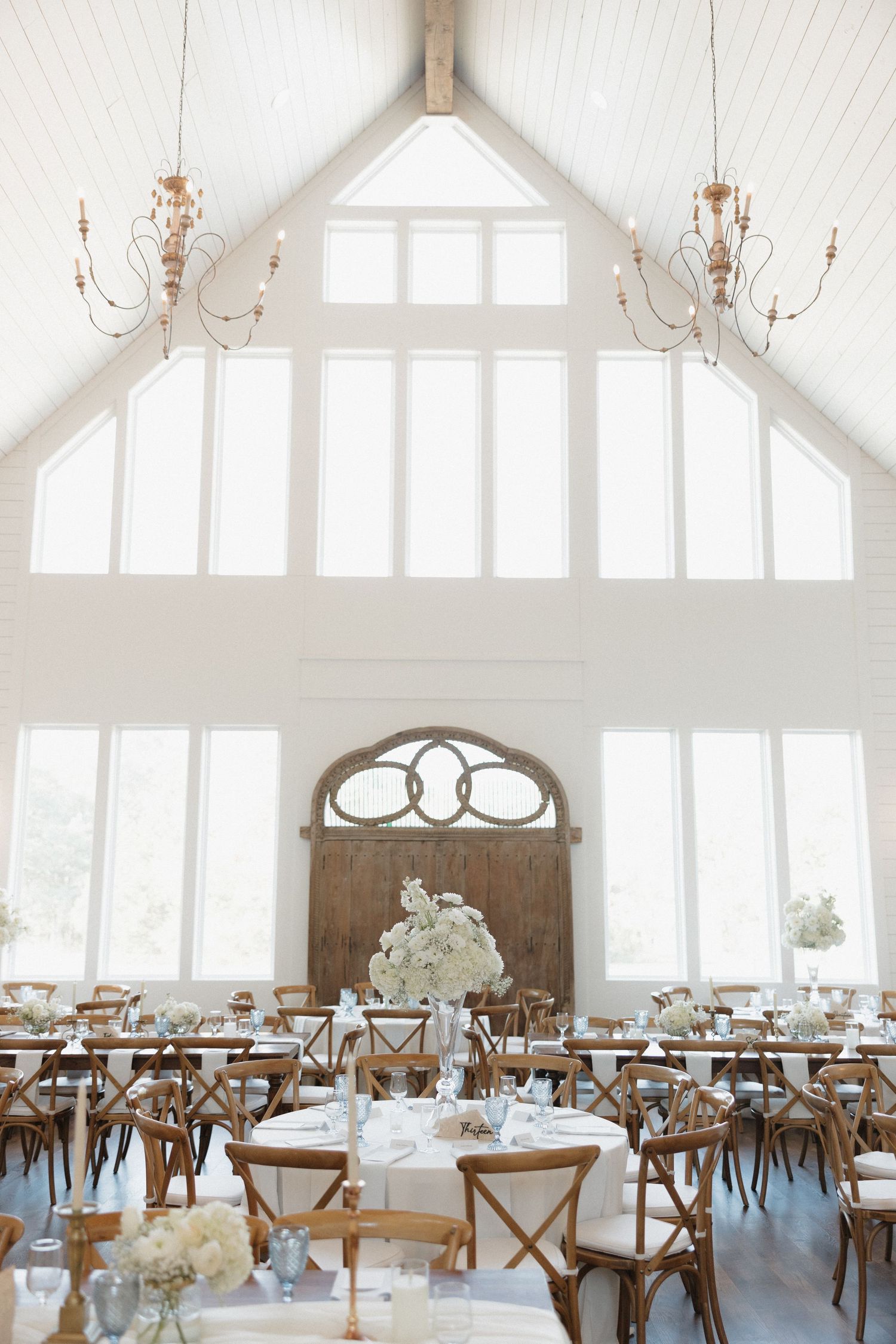 Elegant white wedding reception venue with rustic wood tables, chandeliers, and vaulted ceilings creating an airy atmosphere.