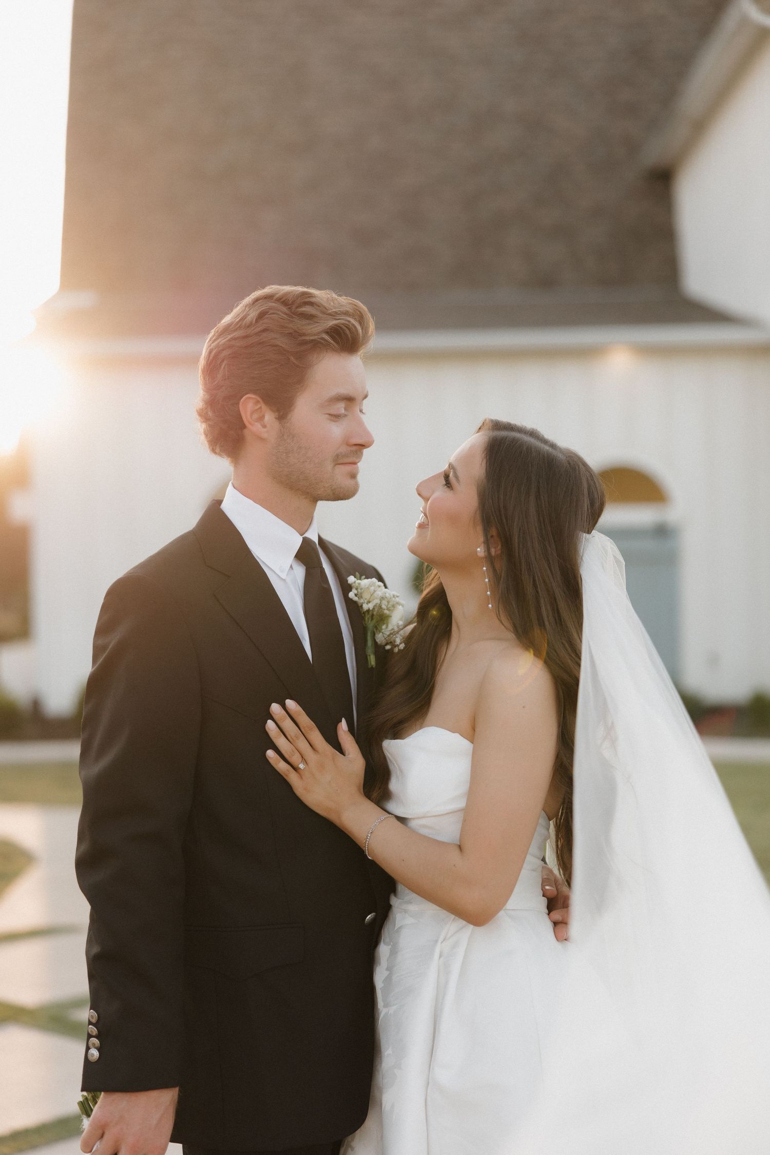 A bride and groom share an intimate moment at sunset outside a white church building.