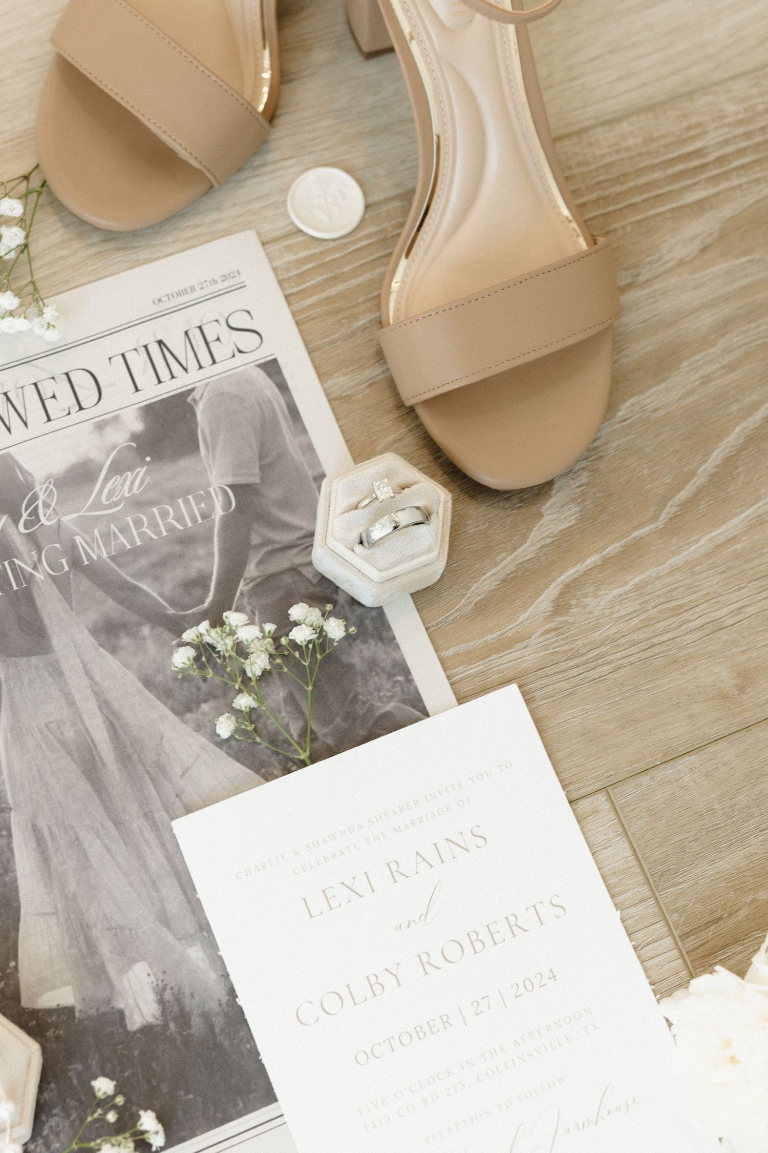 Wedding invitation suite with neutral beige sandals and delicate white flowers arranged on light wooden surface.