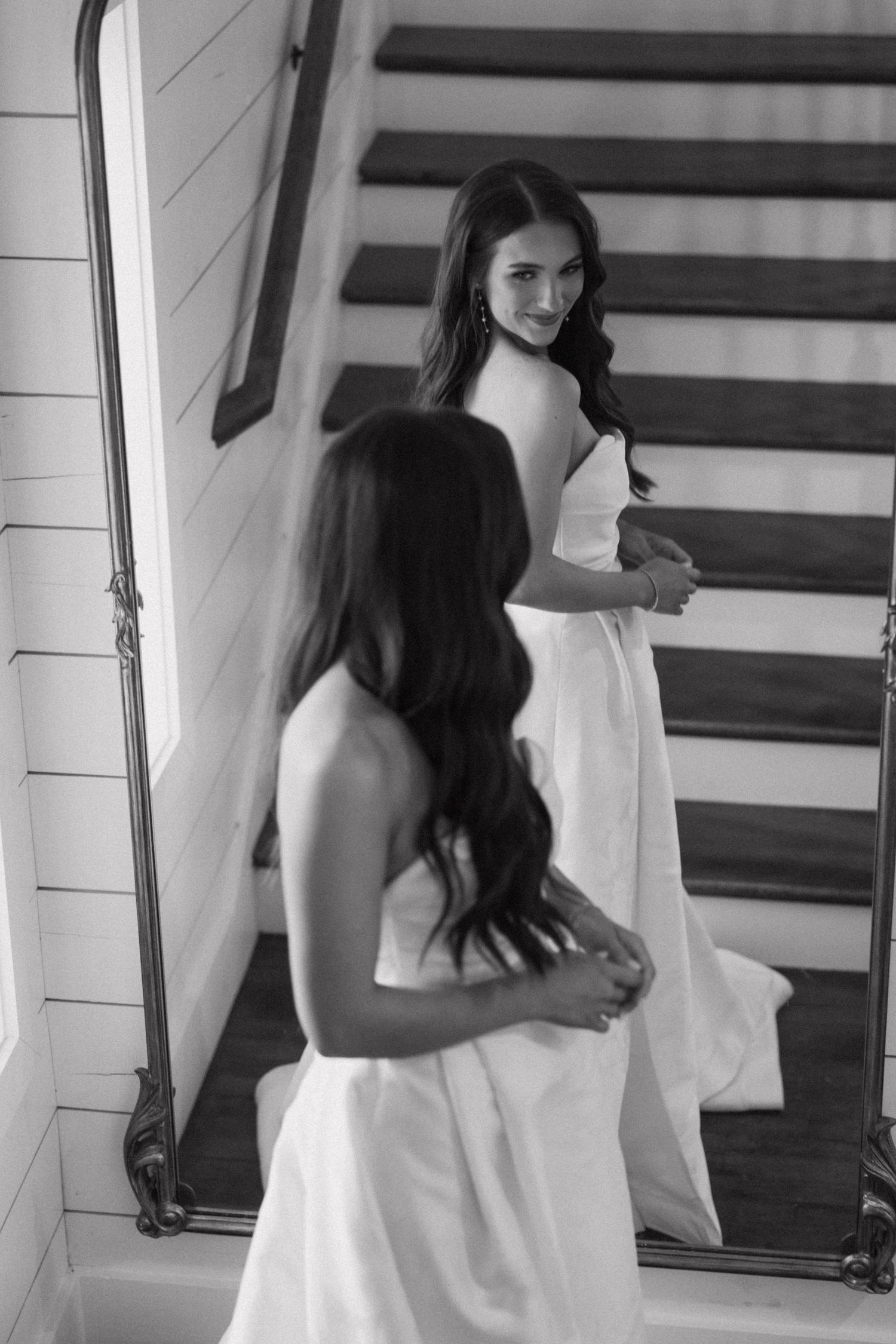 Black and white photo of someone in a white dress looking at their reflection in a mirror by a staircase.