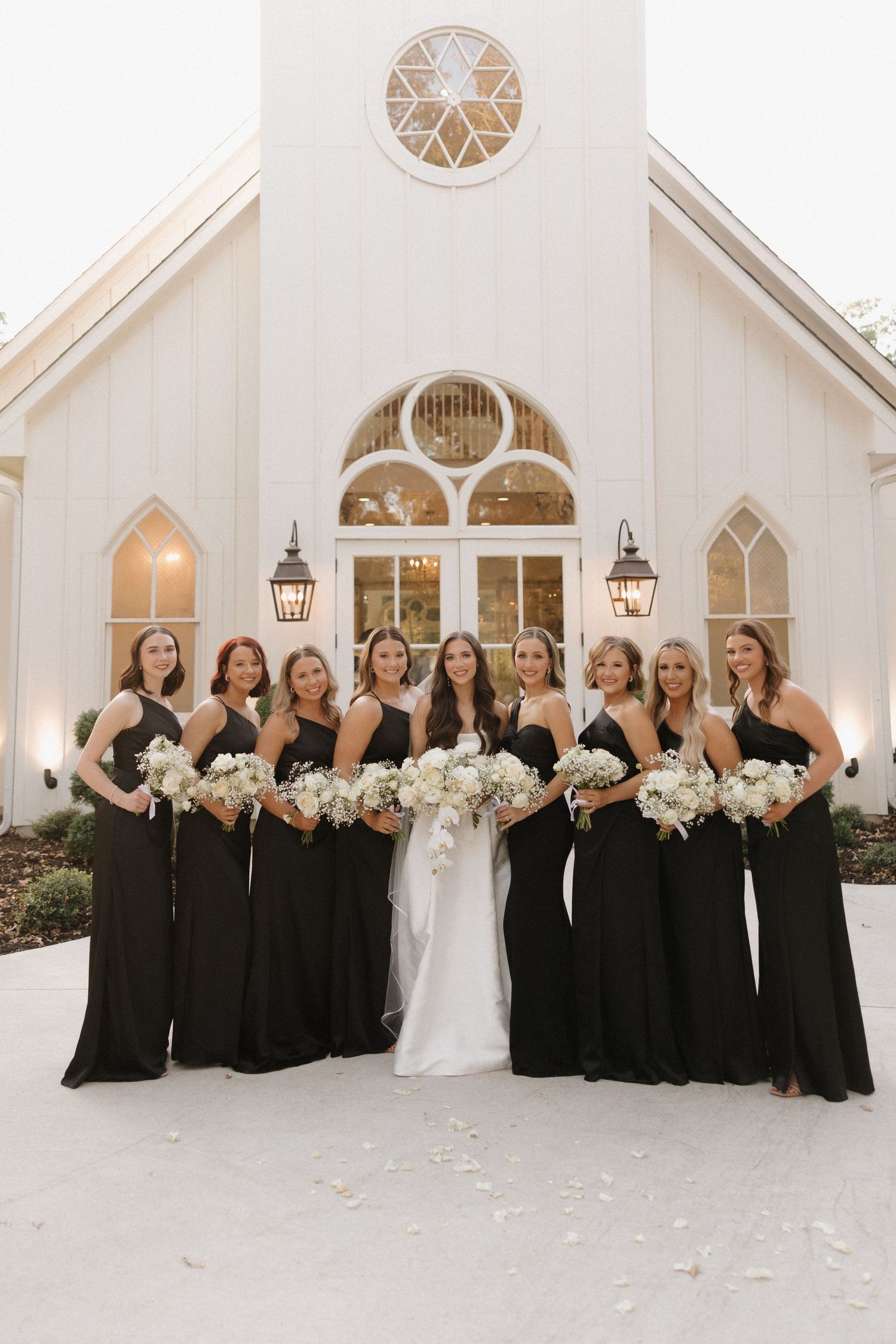 Wedding party in black dresses and white gown pose in front of elegant white chapel with arched windows.