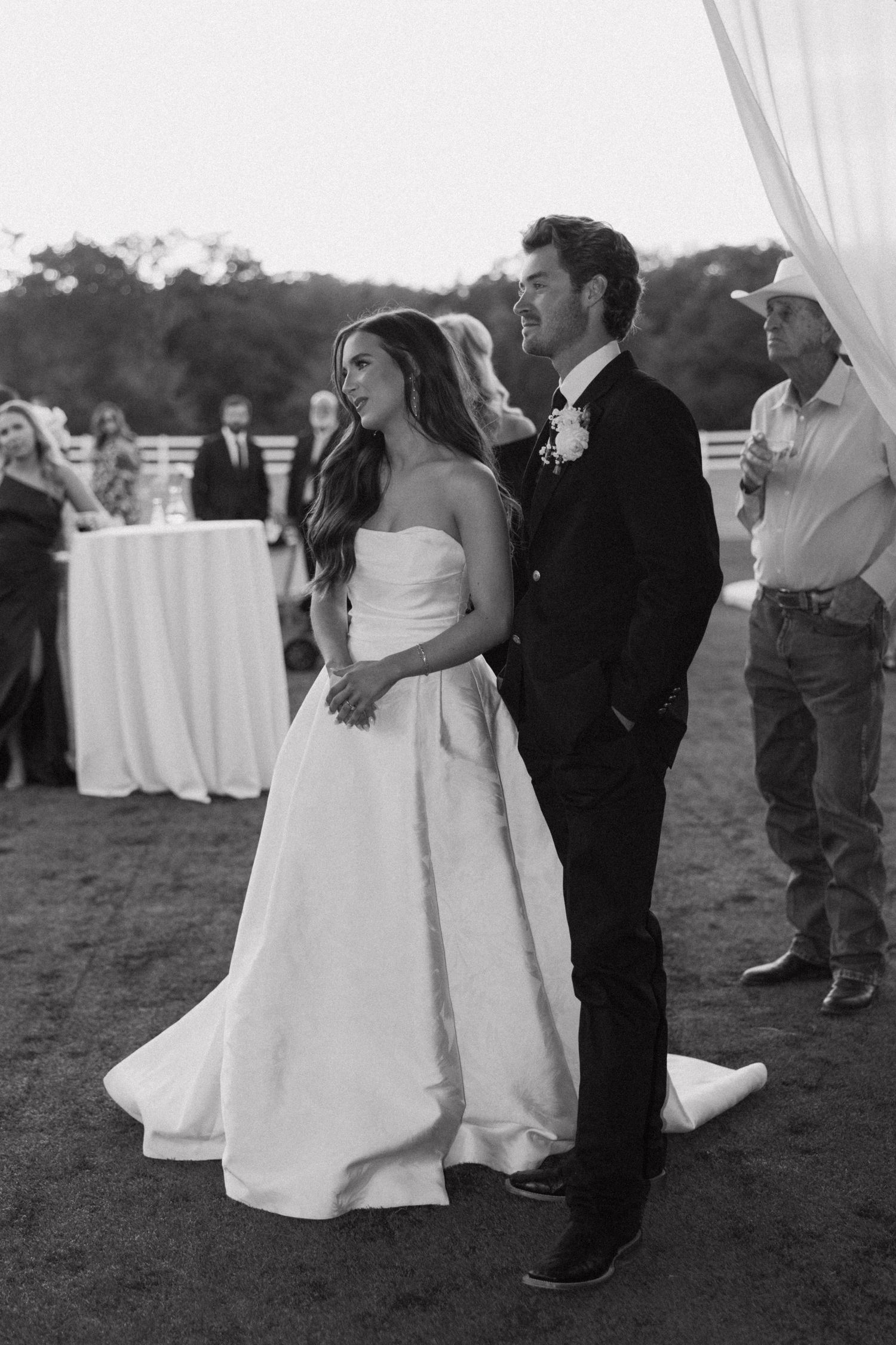 A couple shares a tender moment on their wedding day in black and white as guests mingle in the background.