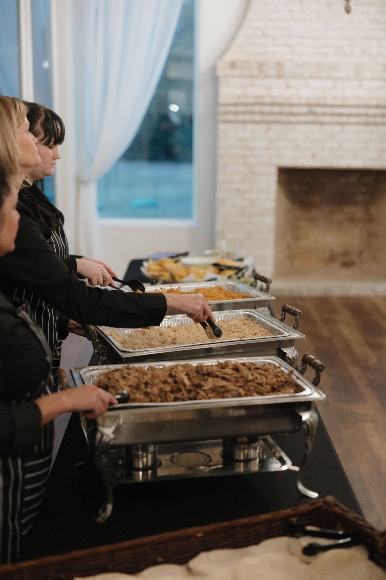 Buffet line with chafing dishes of food being served at an indoor event near a brick fireplace.